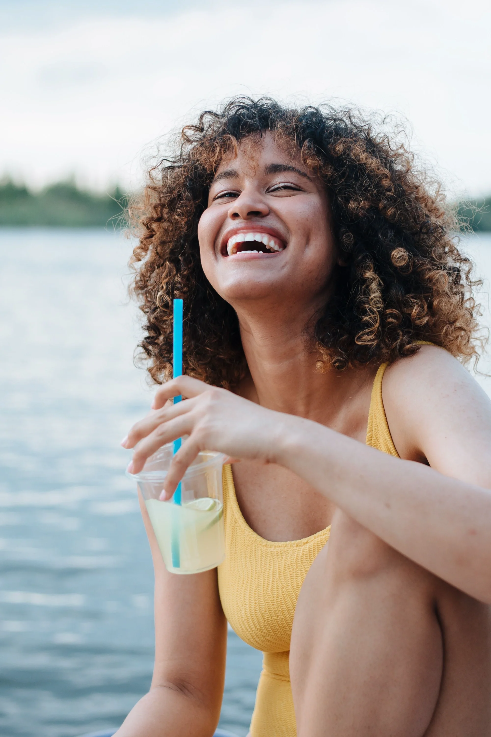 A woman sitting near a lake sipping on a cup of lemonade and smiling at the camera.