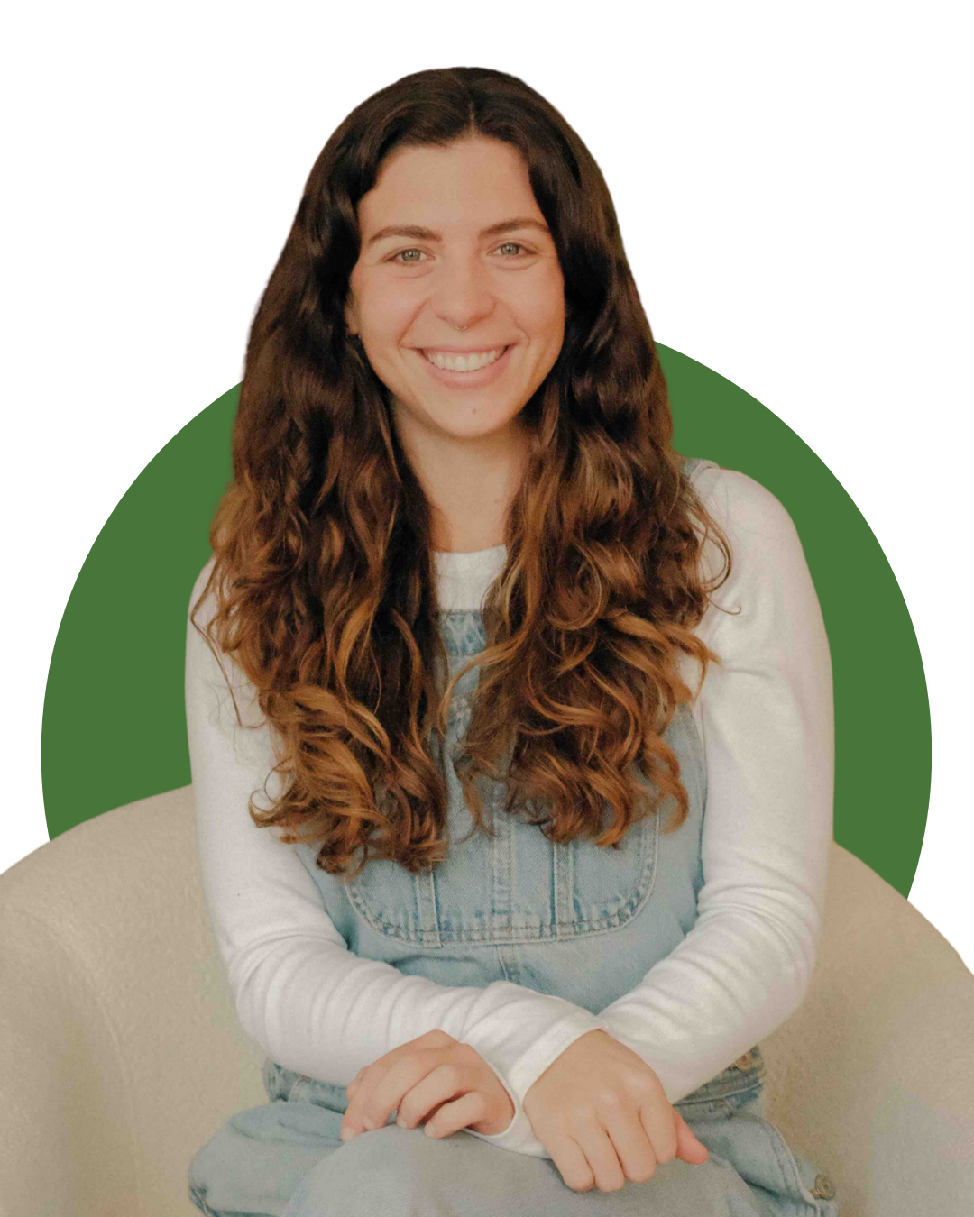  with long, curly brown hair smiling and sitting on a white armchair in a cozy living room. Behind her is a wooden fireplace mantel with decor items, including a framed photo, a small potted plant, and a decorative owl. There are three framed portraits of landscapes or abstract art hanging on the wall above. To the right, there is a large green leafy plant and a small side table with more plants and objects.