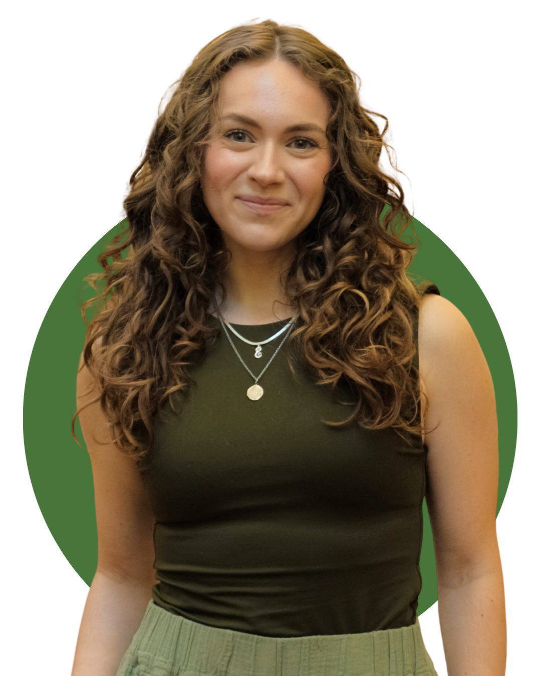 A woman sitting on a beige armchair holding a white mug, smiling, with long curly brown hair, wearing an olive green sleeveless top and pants, in a cozy living room with green plants and framed pictures on the wall.