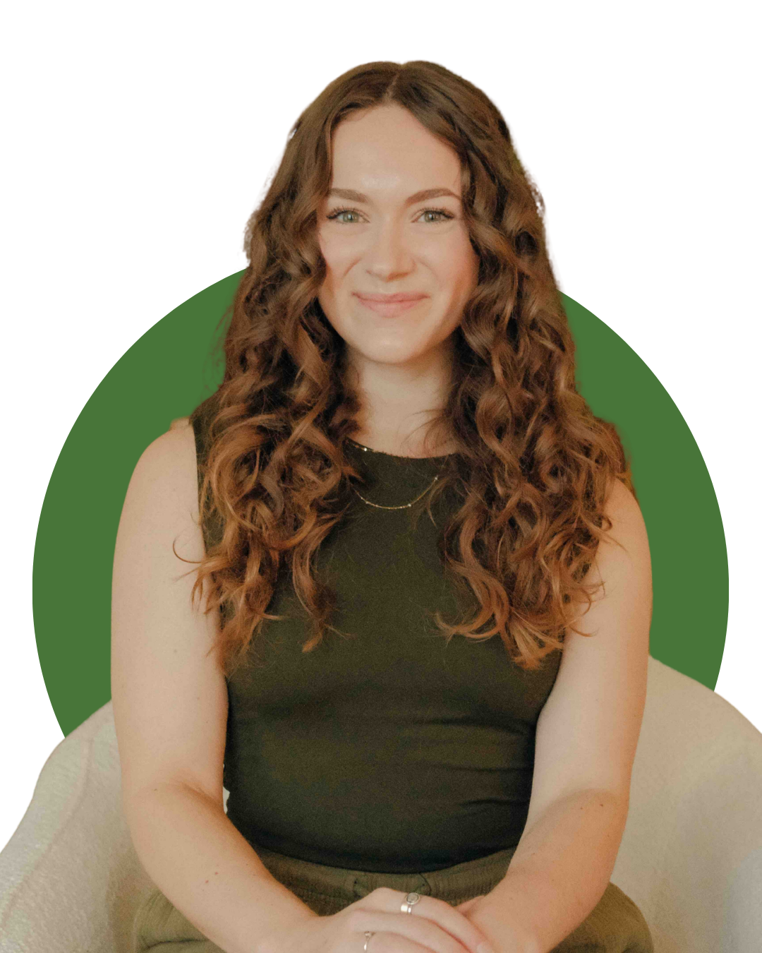 A woman sitting on a beige armchair holding a white mug, smiling, with long curly brown hair, wearing an olive green sleeveless top and pants, in a cozy living room with green plants and framed pictures on the wall.