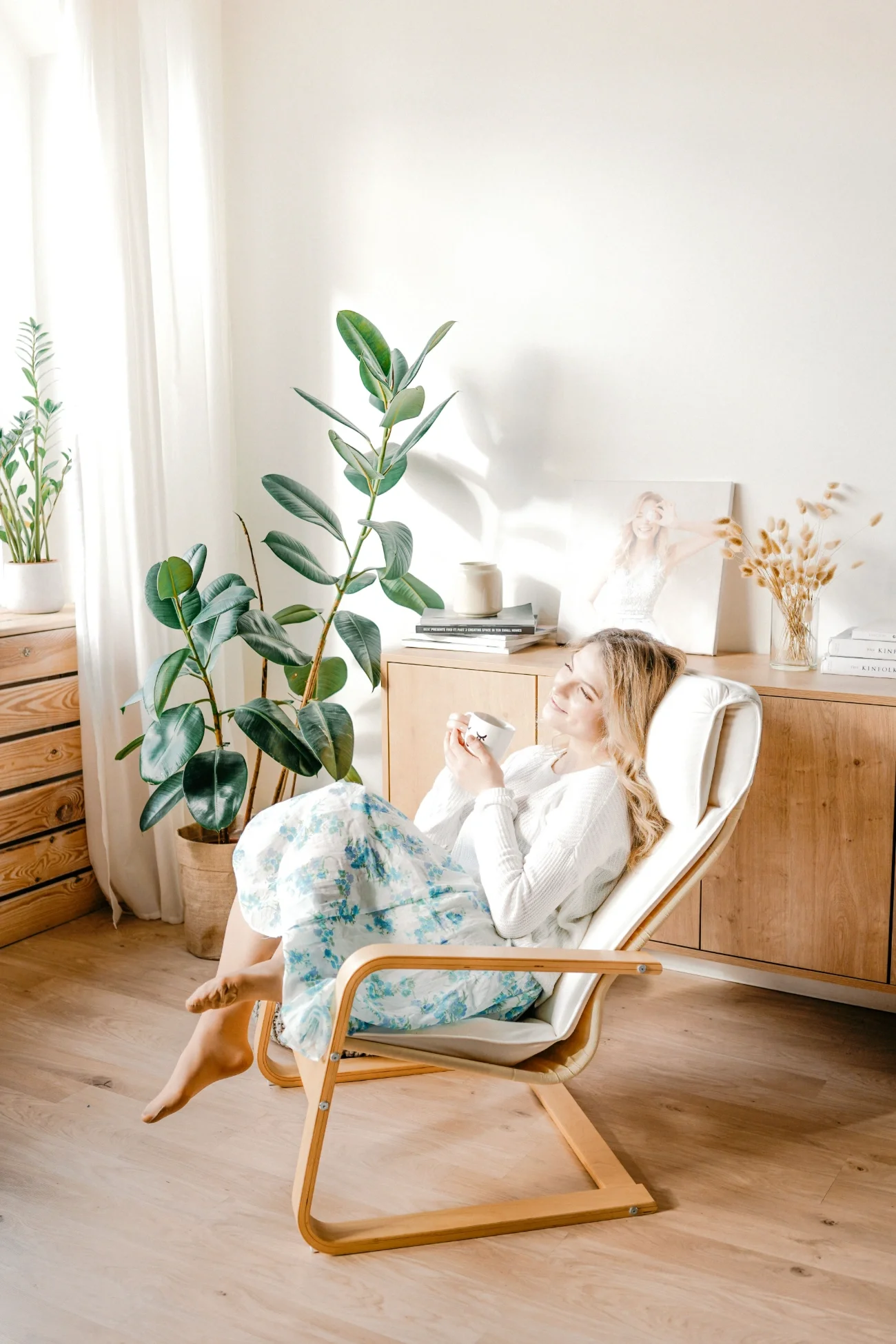 A woman sitting in a chair with a coffee mug in a light coloured room with greenery.