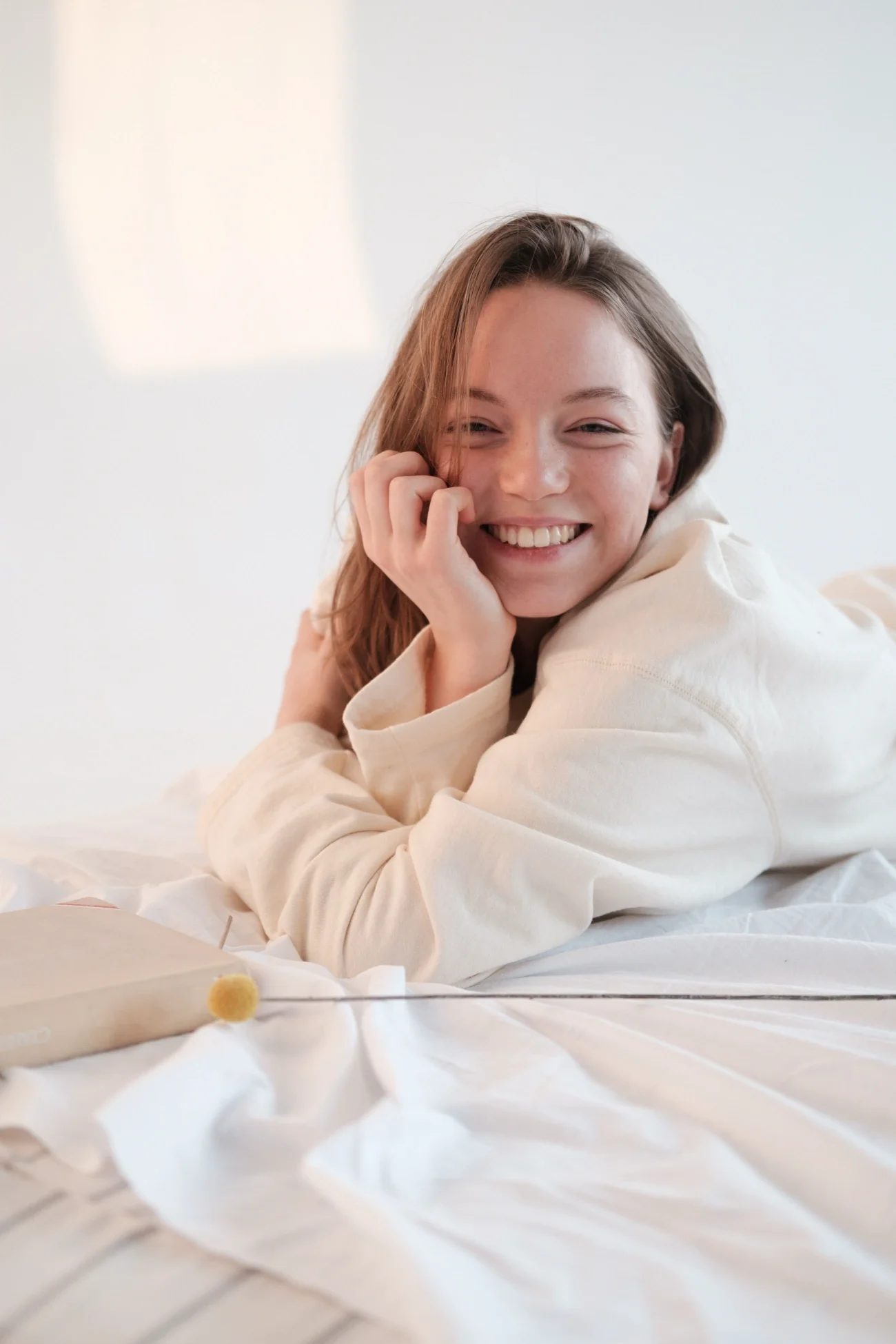 A woman laying on white sheets smiling at the camera with her hand up to her face.