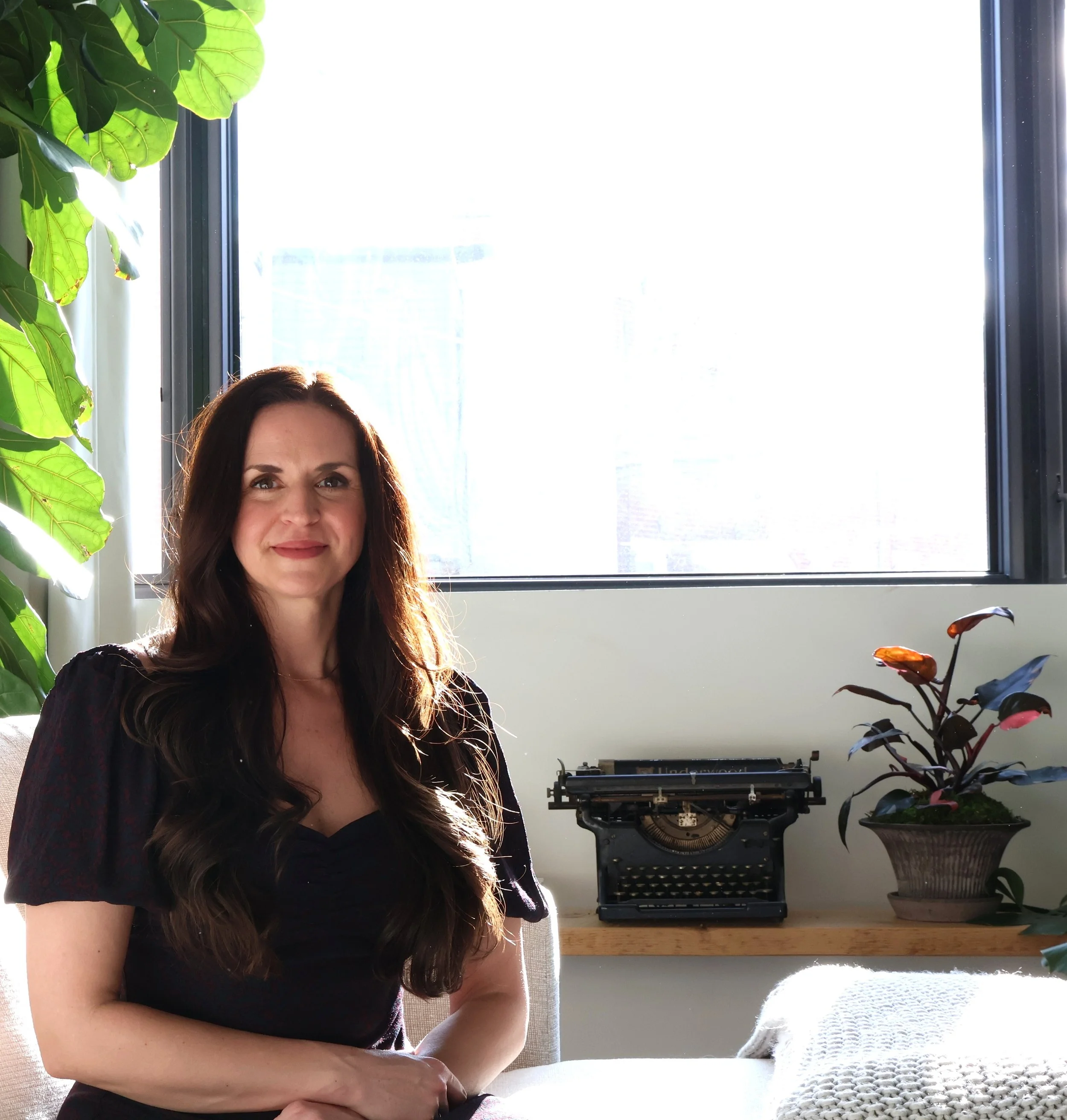 Founder of Hauza Studio, Ashley Hauza, sitting in a room with a large window, green plants, a vintage typewriter on a wooden shelf, and a potted plant with reddish leaves.