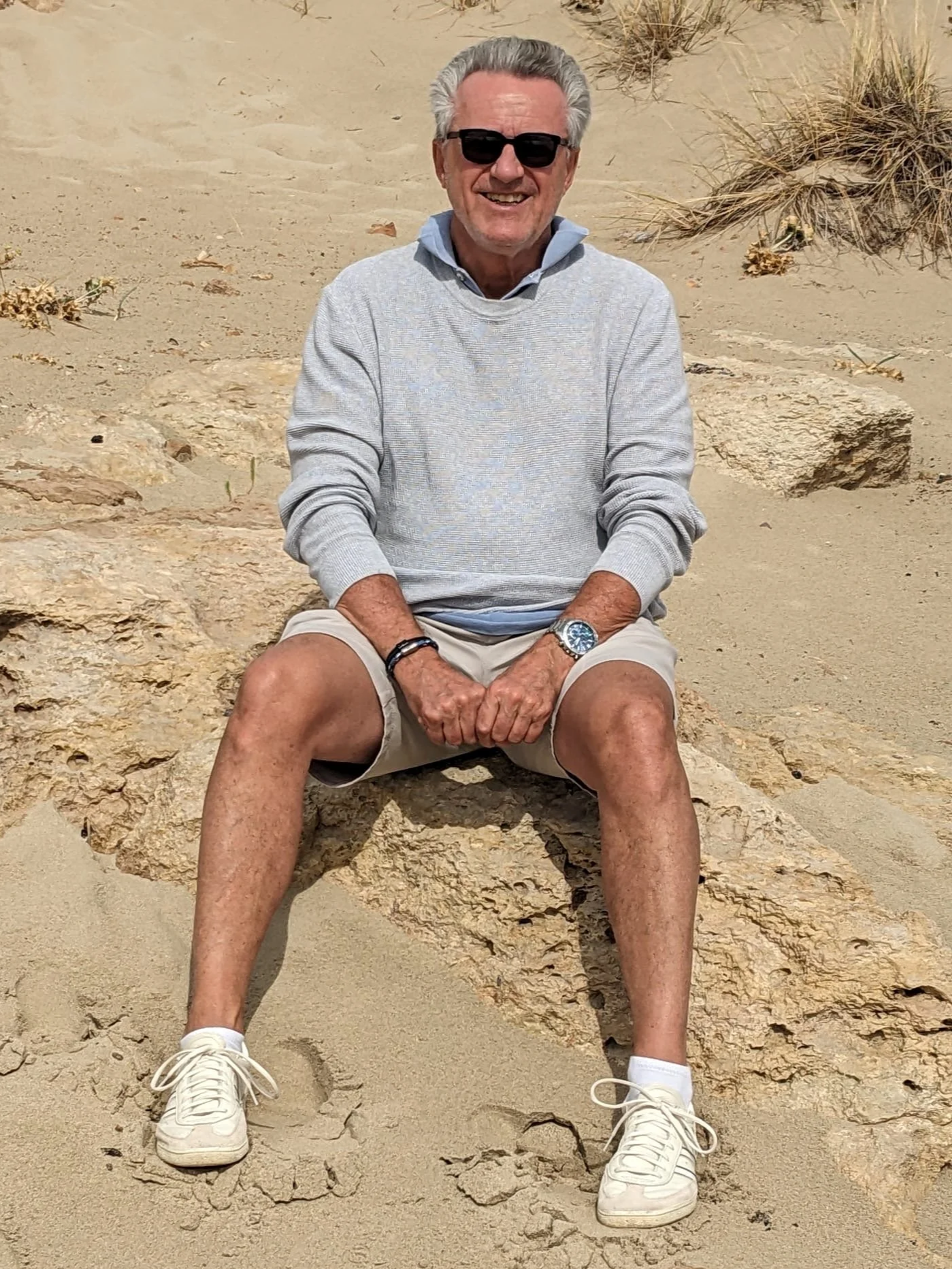 The artist - a man with gray hair and sunglasses, smiling, sitting on a rock in a sandy desert with sparse vegetation, wearing a gray sweater, beige shorts, white sneakers, a watch, and a bracelet.