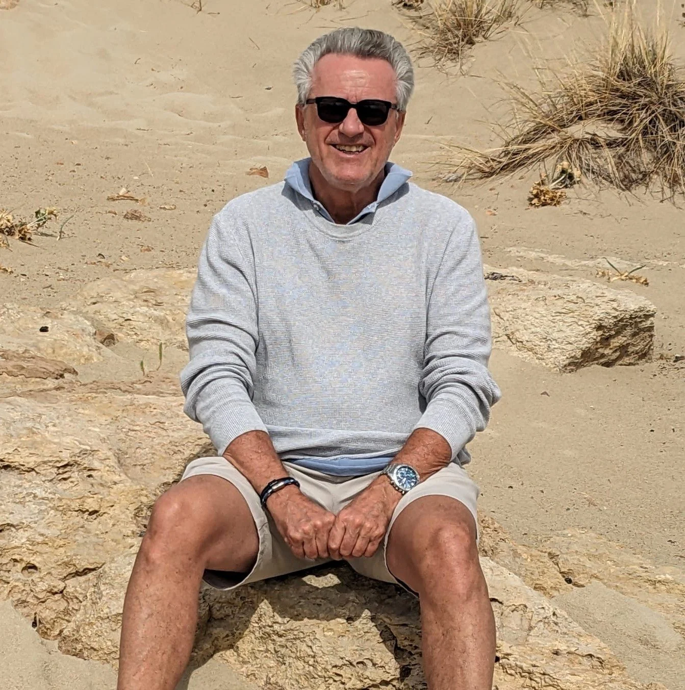 An older man with gray hair, wearing sunglasses, a light gray sweater, and shorts, sitting on a rock in a sandy desert area with sparse vegetation, smiling at the camera.