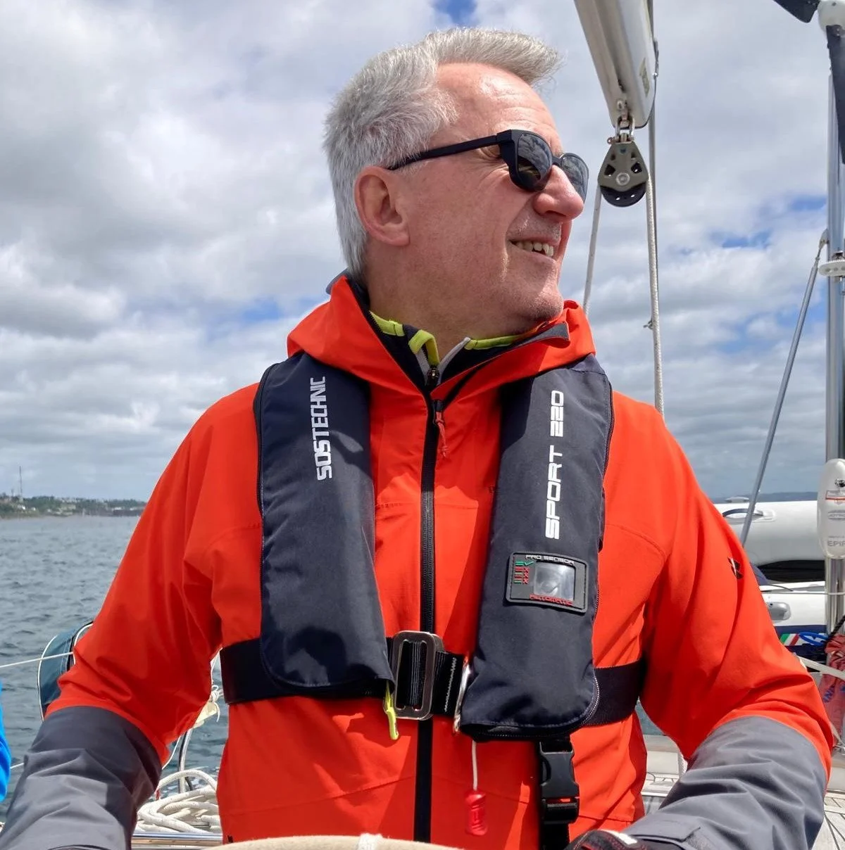 A man wearing sunglasses, an orange weather jacket, and a black life vest smiling on a sailboat during cloudy weather.