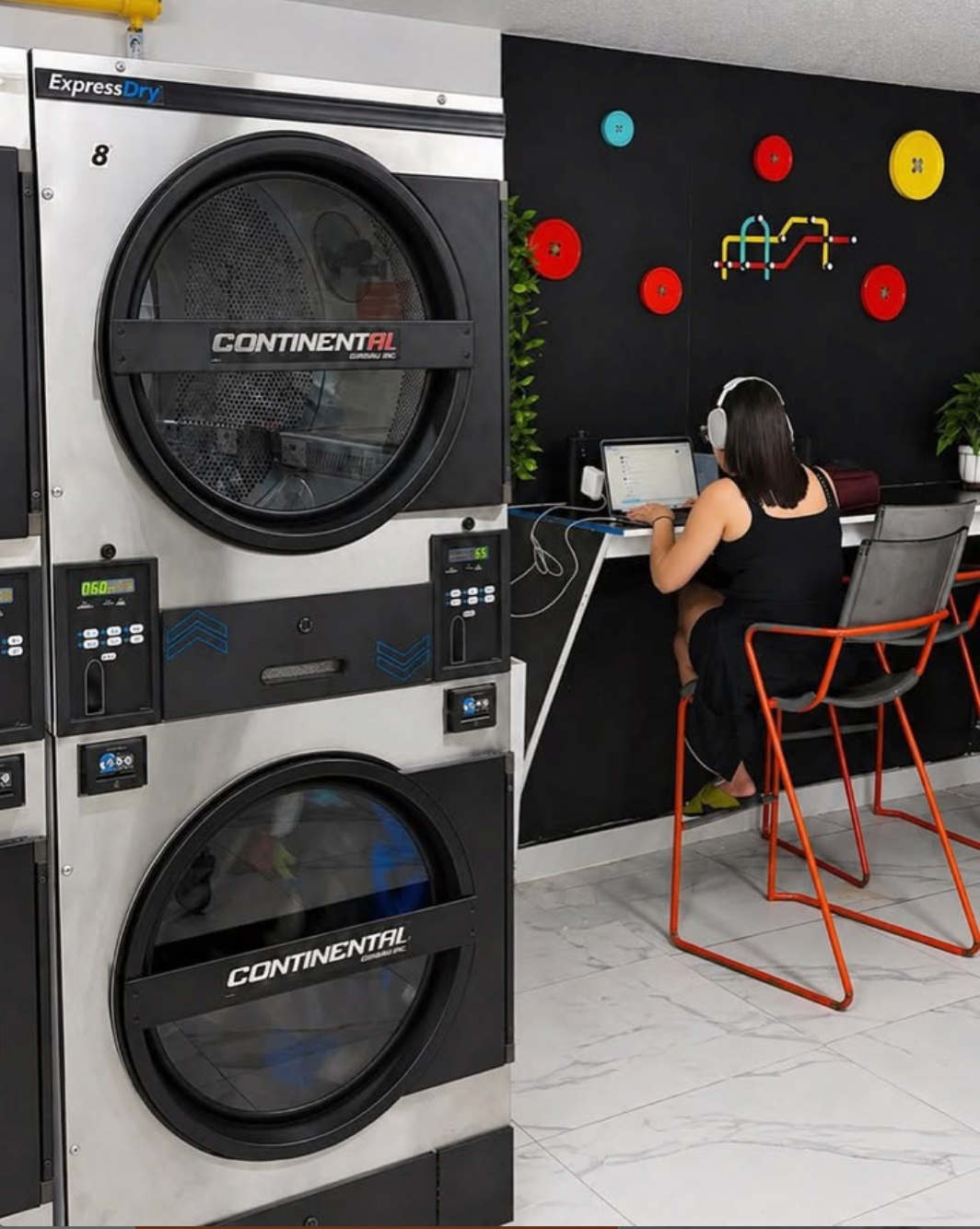 Lavadora y la mujer sentada en una silla con audífonos, trabajando en una computadora portátil en una lavandería moderna con paredes decoradas.