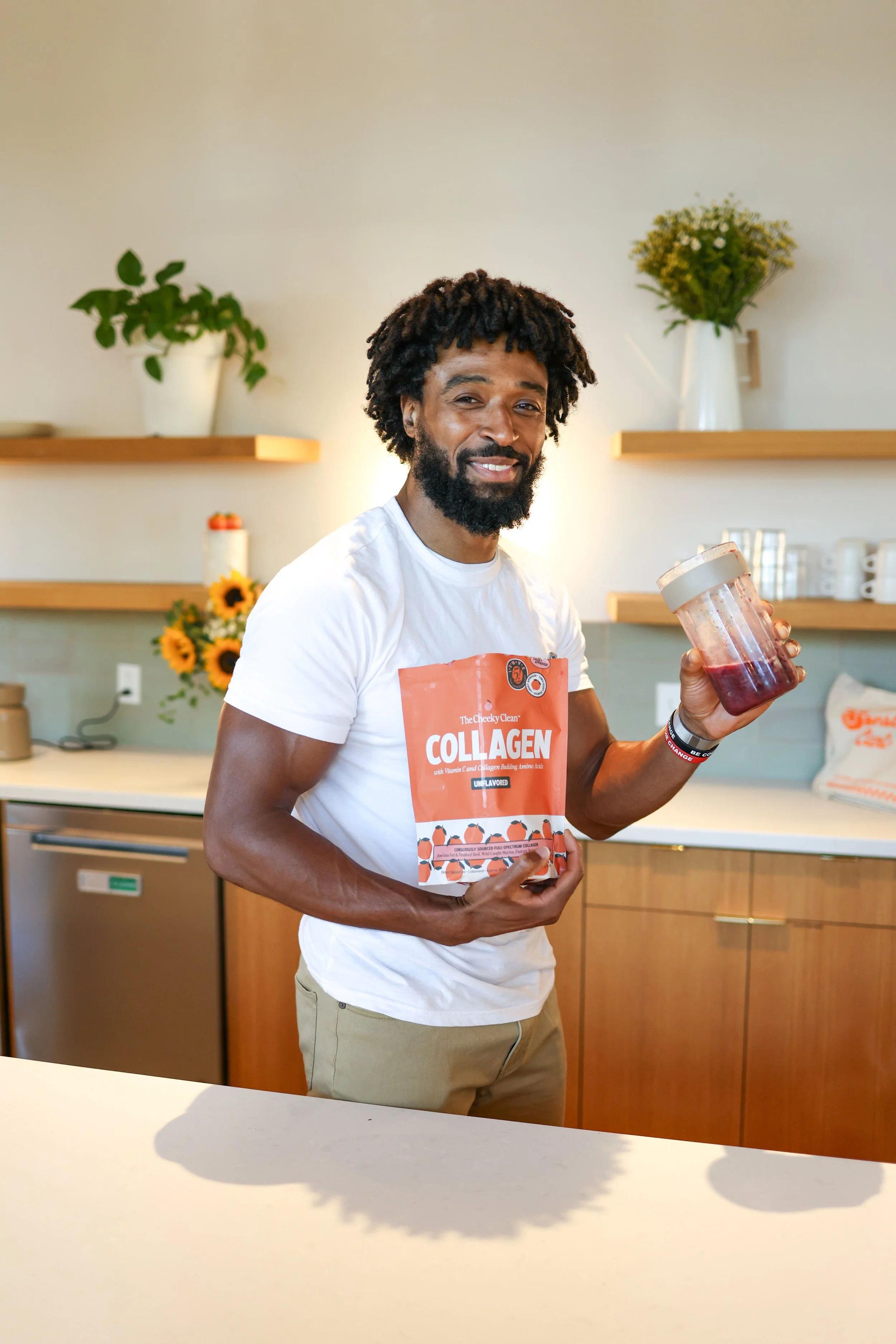 A smiling man with a beard and curly hair holding a jar of red smoothie and a packet of collagen powder in a kitchen with wooden shelves, potted plants, sunflowers, and a counter.