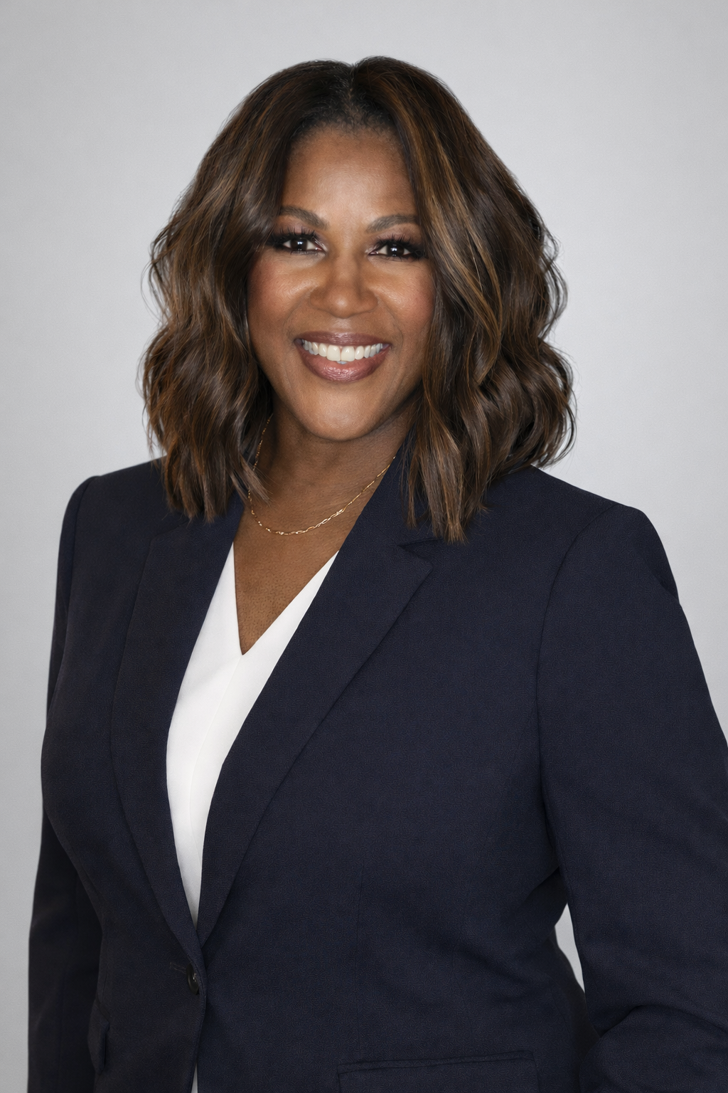 Professional woman with shoulder-length wavy hair wearing a navy blazer over a white top, smiling against a plain light gray background.