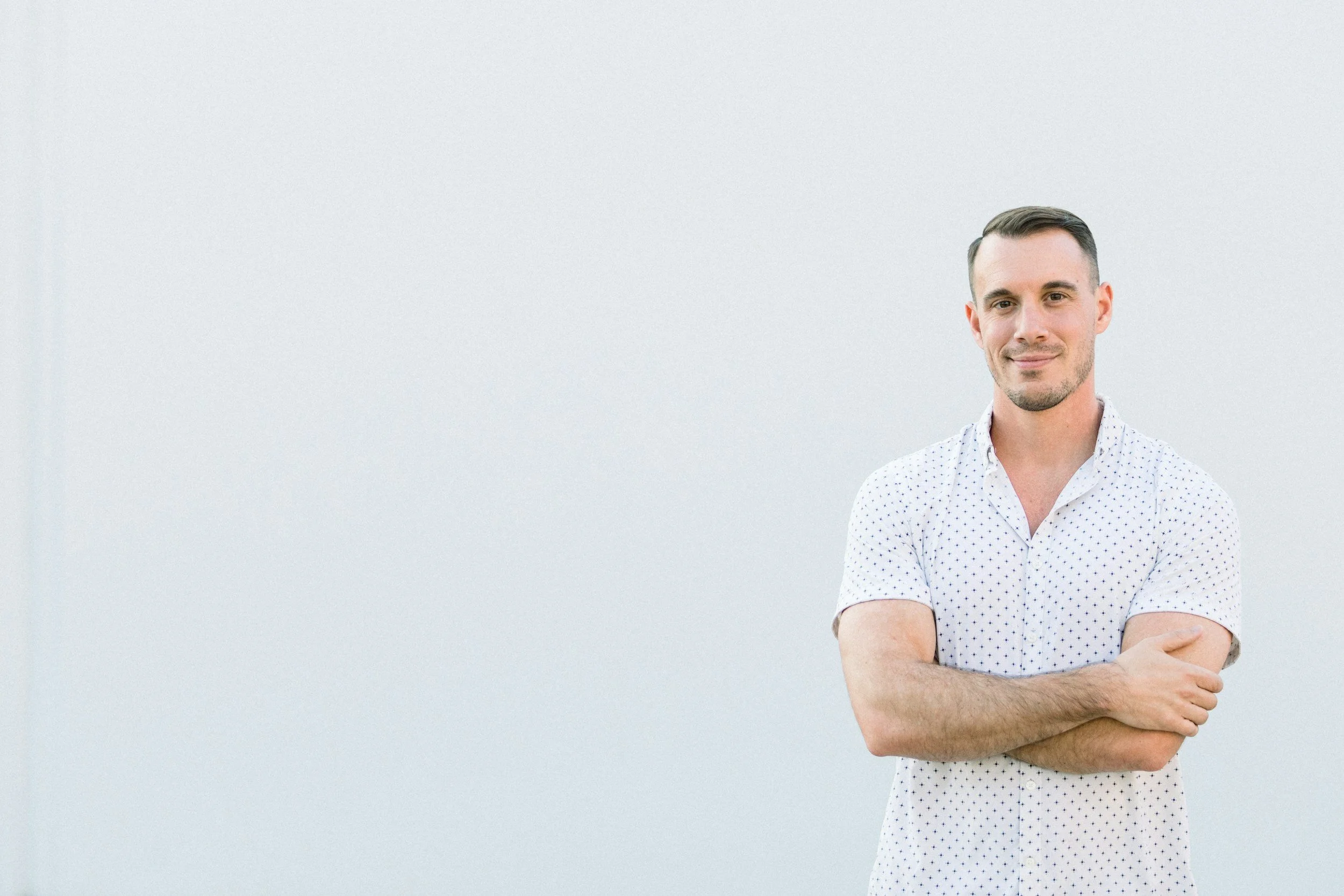 A man standing against a plain white wall, crossing his arms, wearing a white shirt with small dark polka dots, smiling slightly.