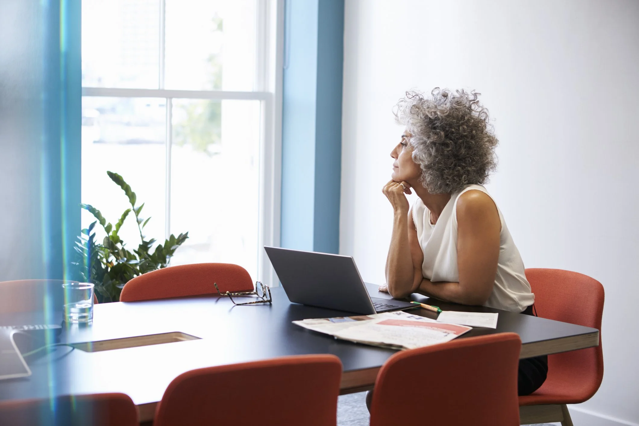 A woman with curly gray hair and a sleeveless white top sitting at a conference table with a laptop, resting her chin on her hand, looking out a window. There are orange chairs, a potted plant, and papers on the table.