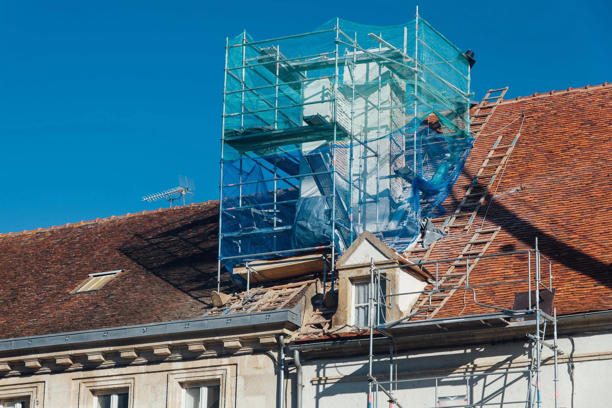 Travaux de rénovation sur le toit d'une maison, avec de l'échafaudage en bleu et une échelle en bois sur le côté.