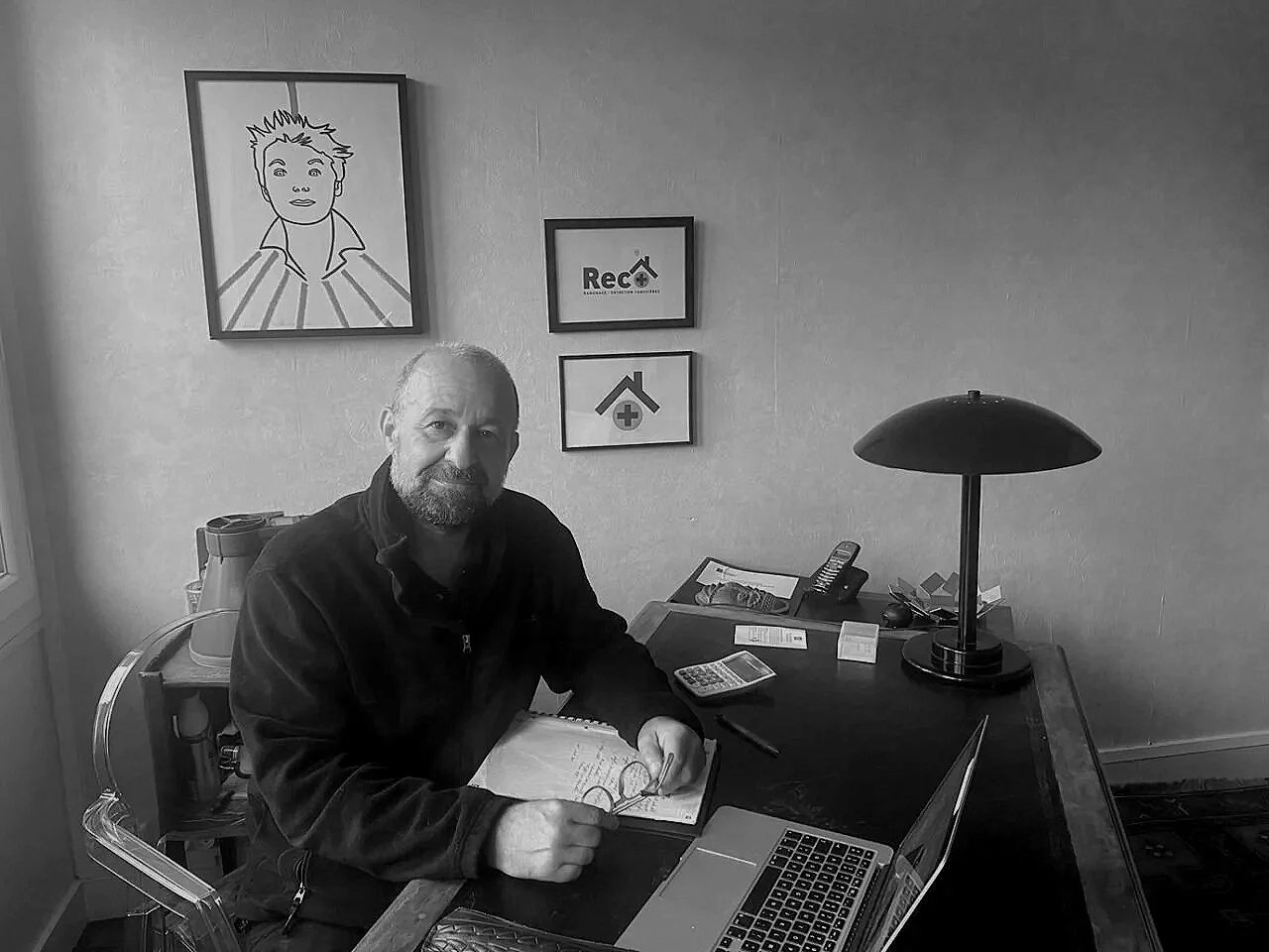 Un homme assis à un bureau avec un ordinateur portable, une lampe, un téléphone, une calculatrice, et des papiers, dans une pièce avec des affiches au mur, dont une avec un dessin d'un garçon et une autre avec le logo RECAT.