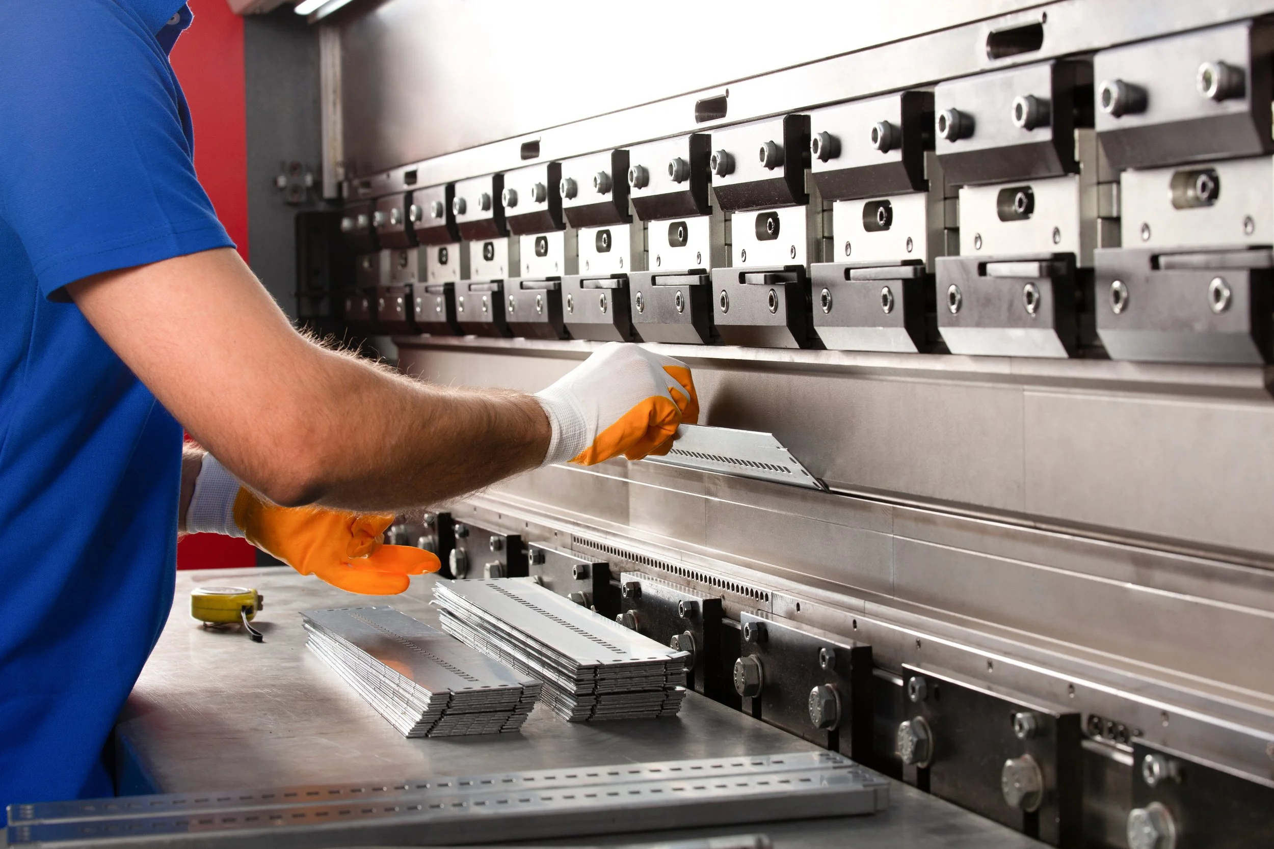 A worker wearing an orange glove and a blue shirt is operating machinery that produces multiple identical metal parts stacked on a worktable.