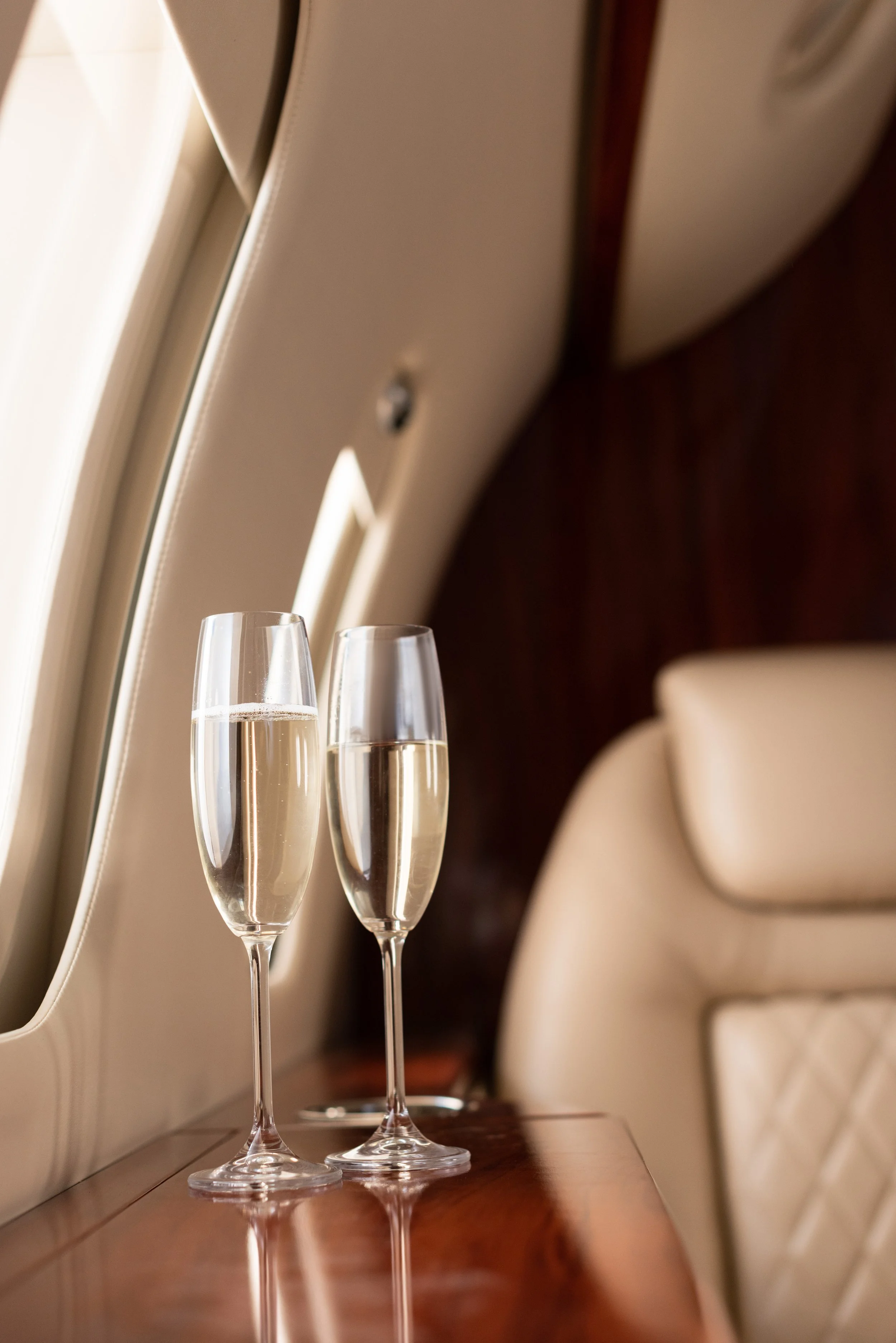 Dos copas de cristal con champagne en un avión, en una mesa de madera junto a una ventana y con asientos de cuero beige en el fondo.