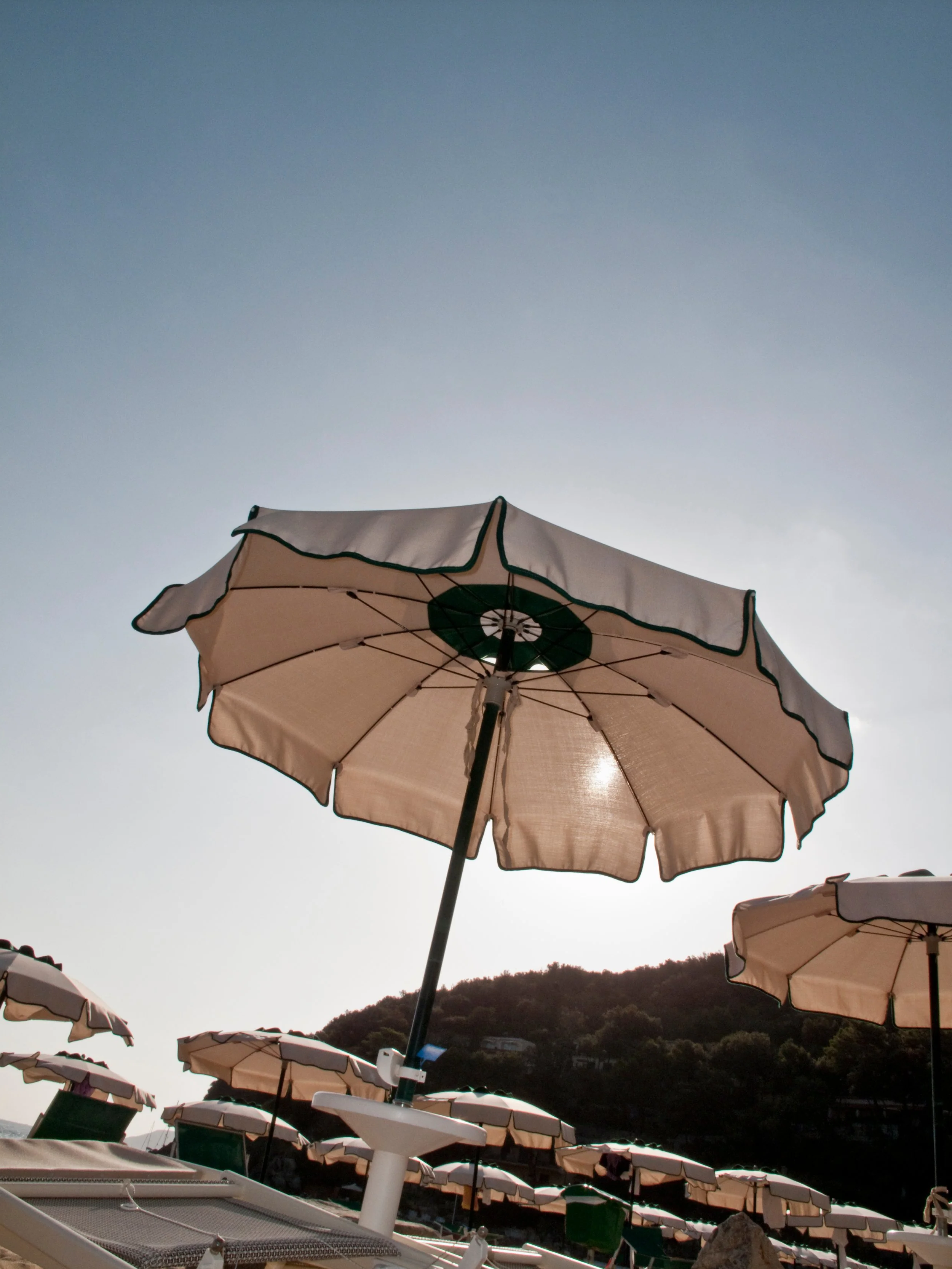 Sombrilla beige con borde oscuro en una playa soleada, con muchas sombrillas similares y colinas verdes al fondo.