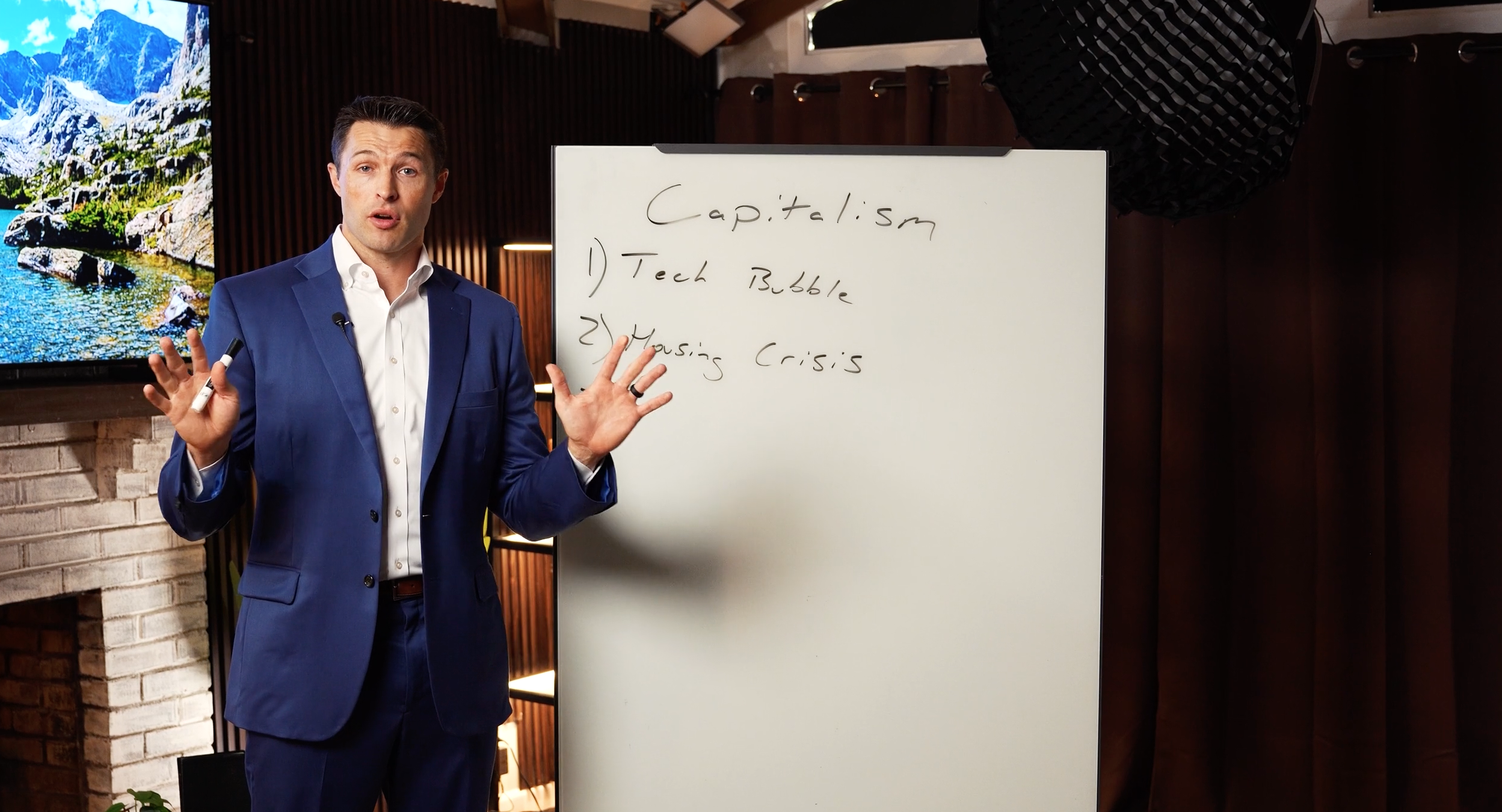 A man in a blue suit giving a presentation with a whiteboard that has notes about capitalism, including topics on tech bubbles and housing crises, in a room with a TV screen displaying a mountain landscape.
