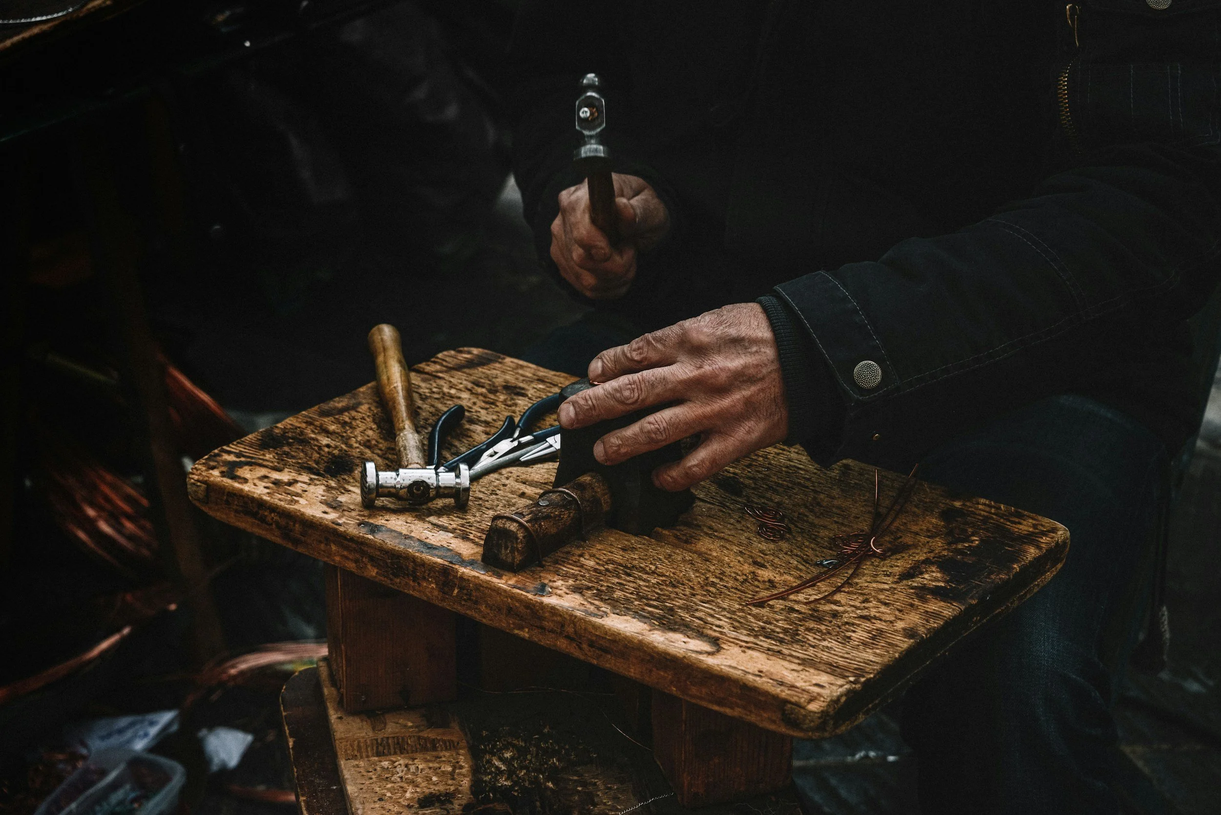 A person using a hammer and pliers at a wooden workbench, with a wire and tools nearby, working on a crafting project.