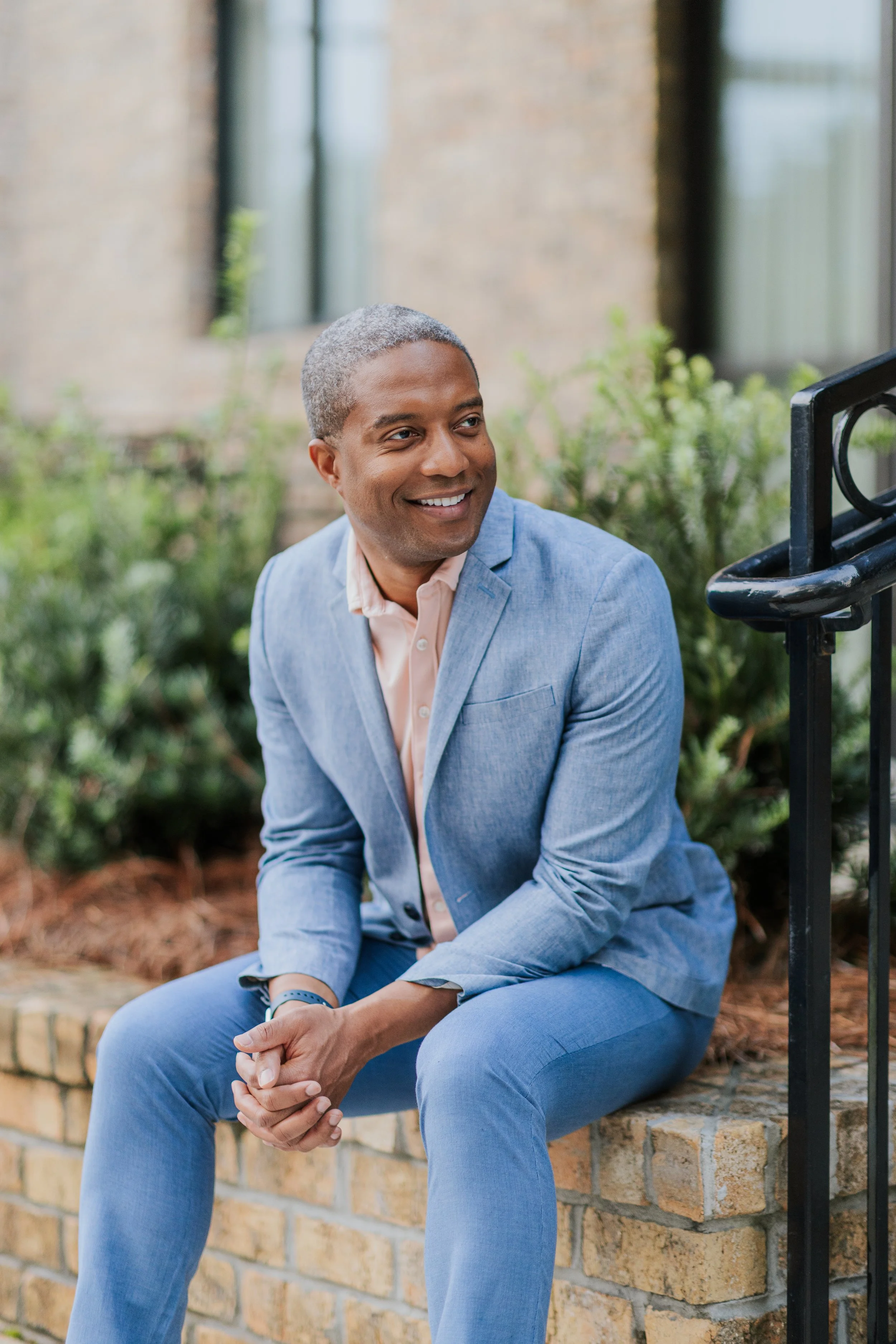A smiling man in a light blue blazer and matching pants sitting on a brick ledge outdoors.
