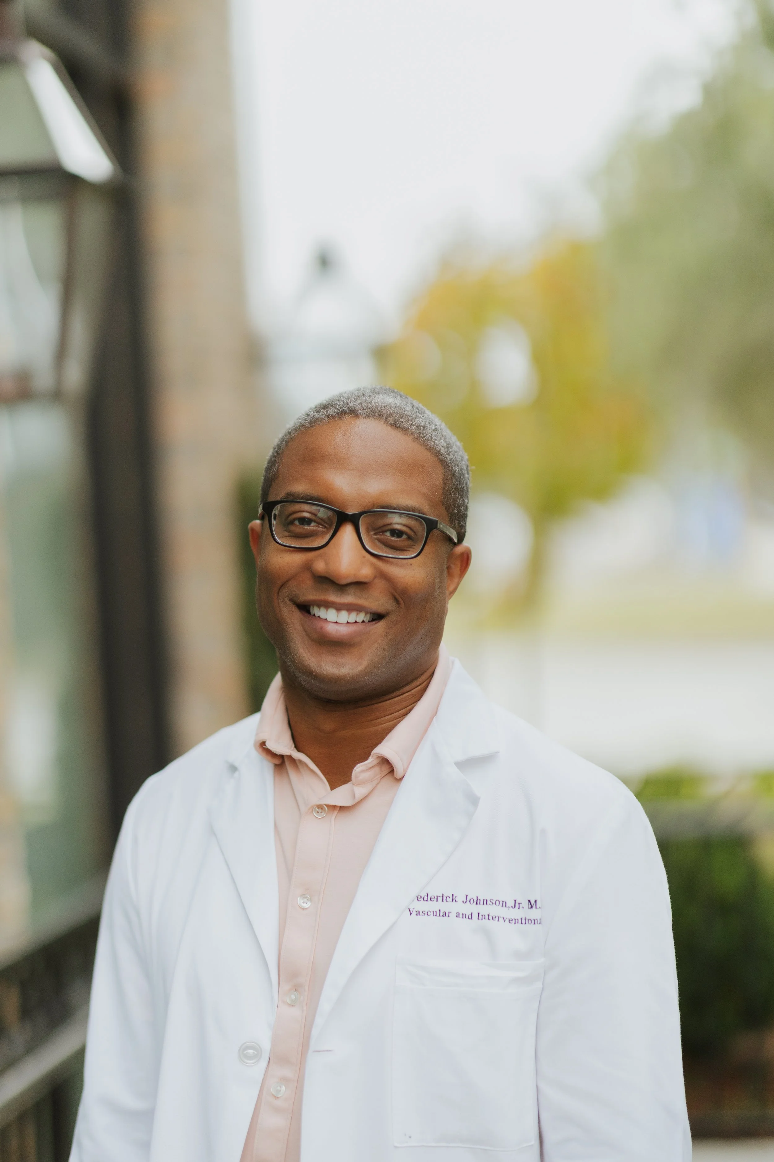 Smiling African American male doctor wearing glasses, a white lab coat, and a light pink shirt standing outside during daytime.