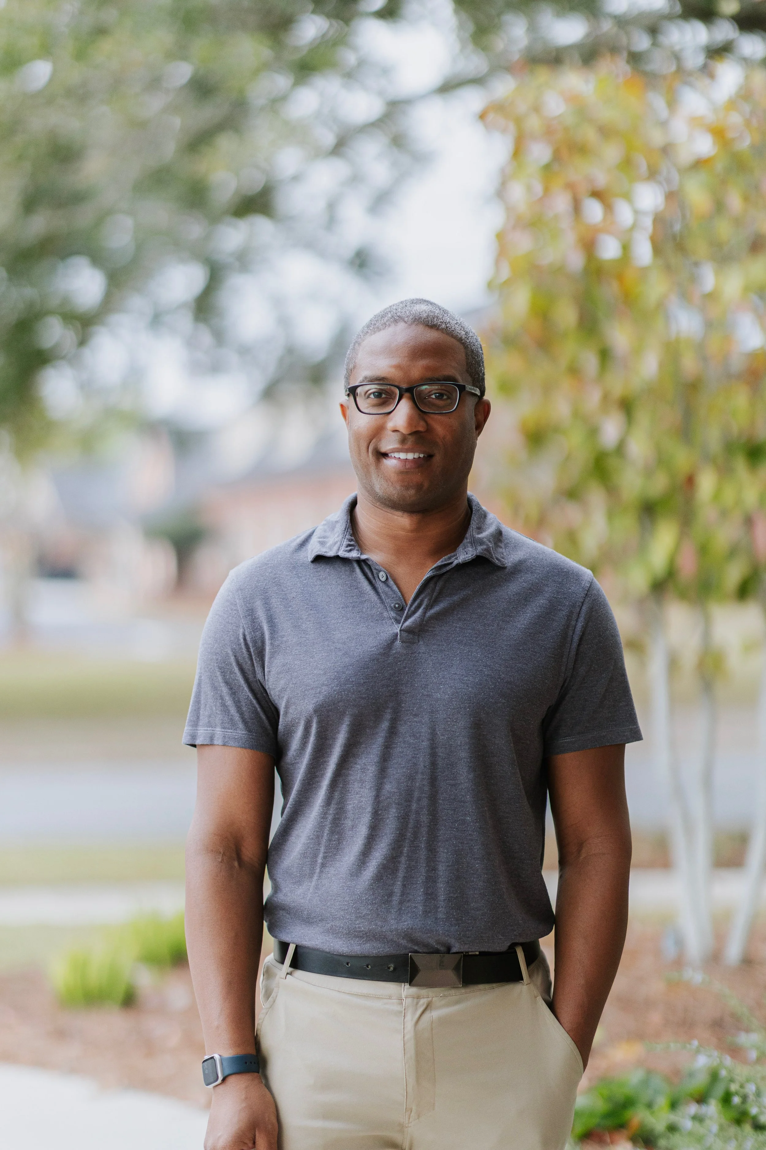 A man with short gray hair, glasses, wearing a dark gray polo shirt and khaki pants, standing outdoors in a natural setting with trees and bushes in the background, smiling at the camera.