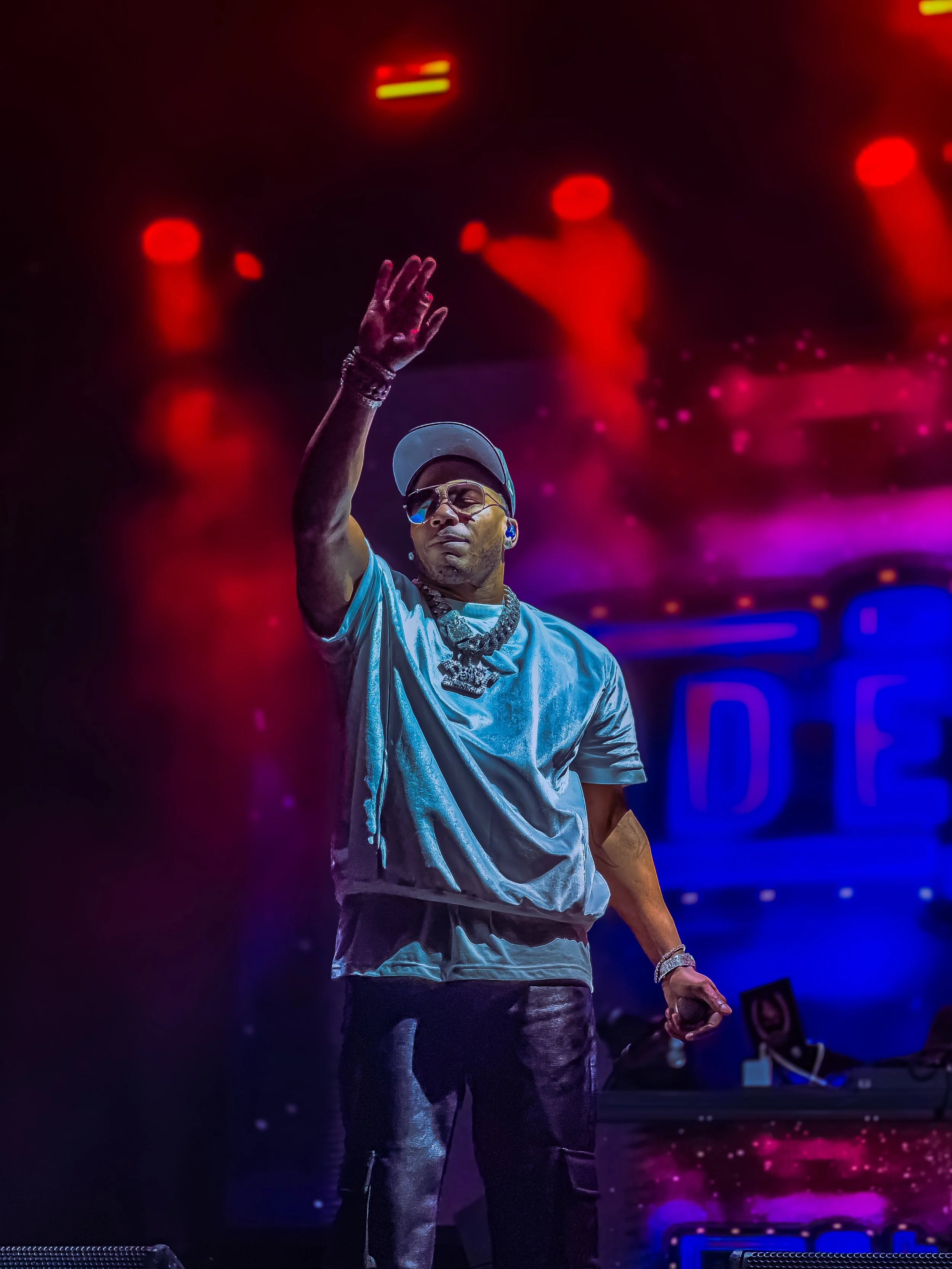 The male rapper Nelly on stage, wearing a shiny silver t-shirt, black leather pants, a cap, sunglasses, and jewelry, performing during a concert with colorful stage lights at ONE Musicfest