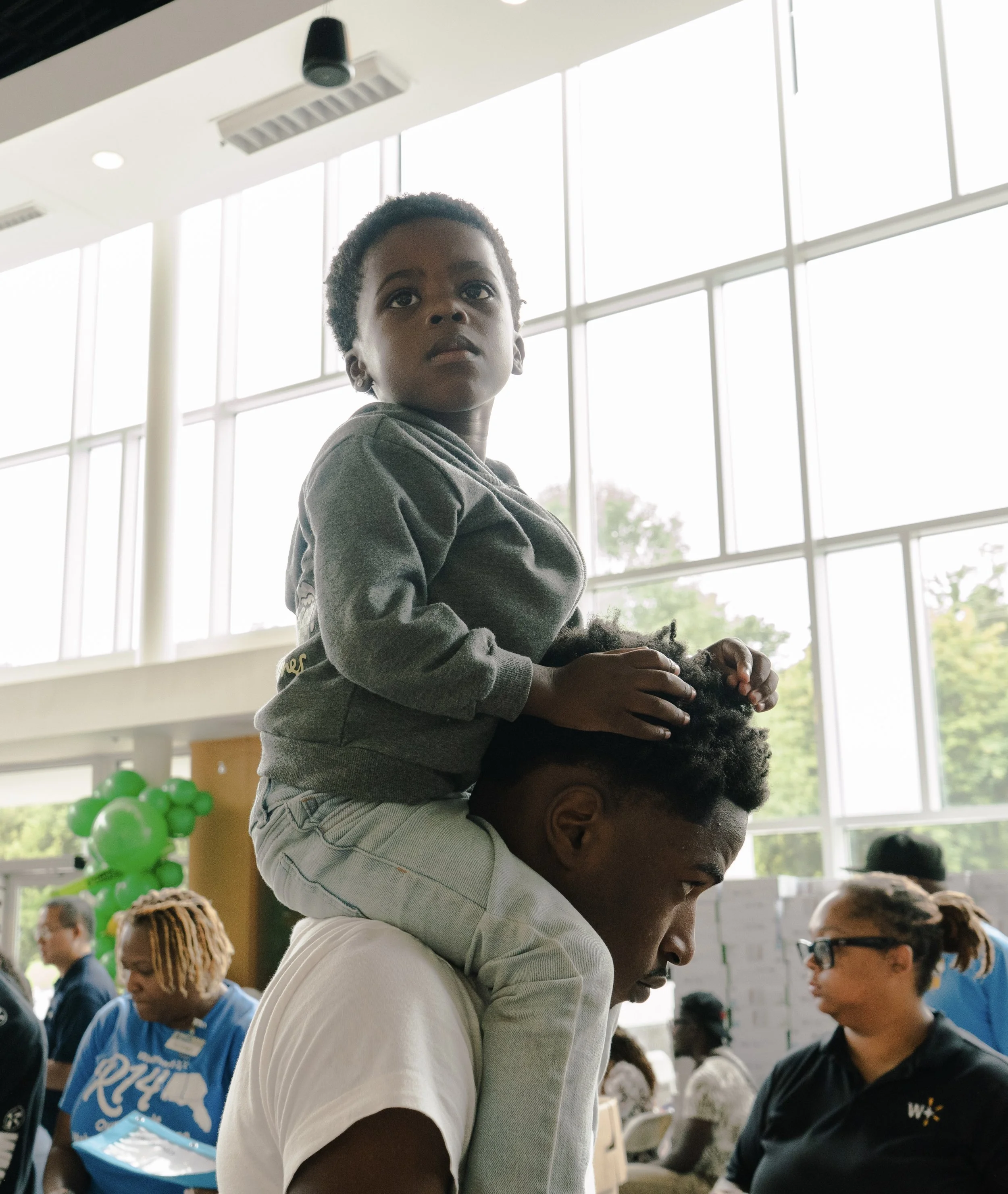A young boy is sitting on a man's shoulders inside a well-lit building with large windows. Several people are in the background, some looking at papers or talking.