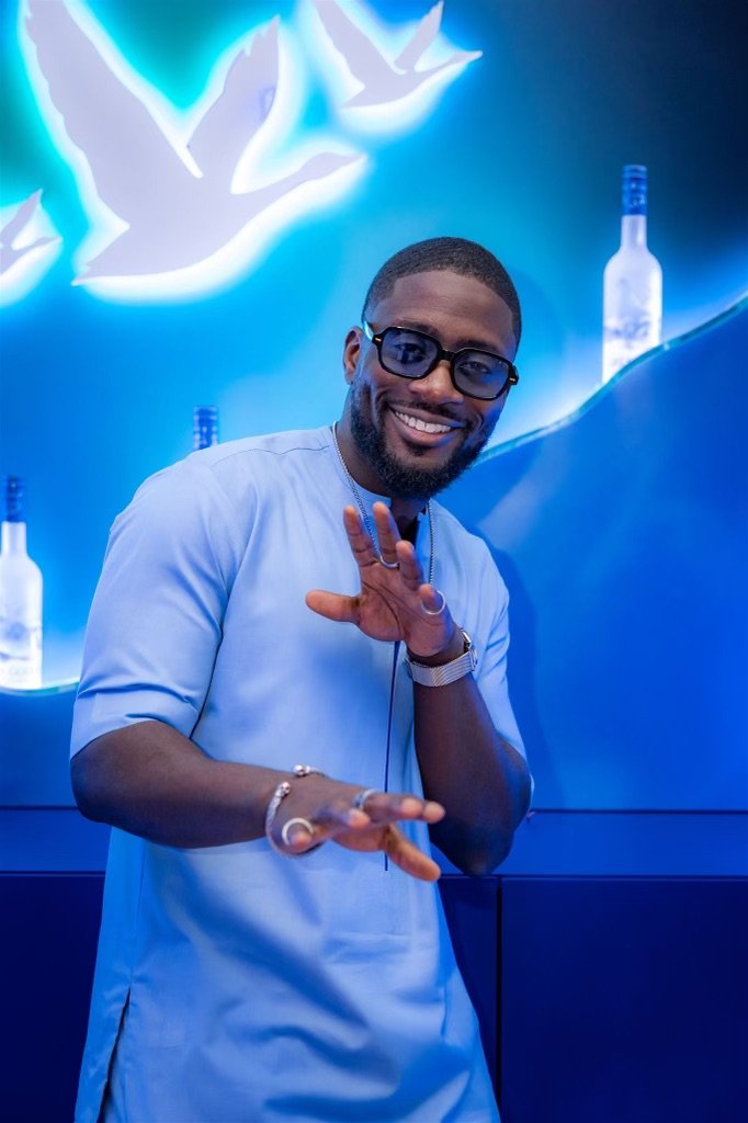 A smiling man with beard and glasses wearing a light blue shirt, posing with hands in a playful gesture, standing against a blue illuminated background with bottles and a bird graphic.