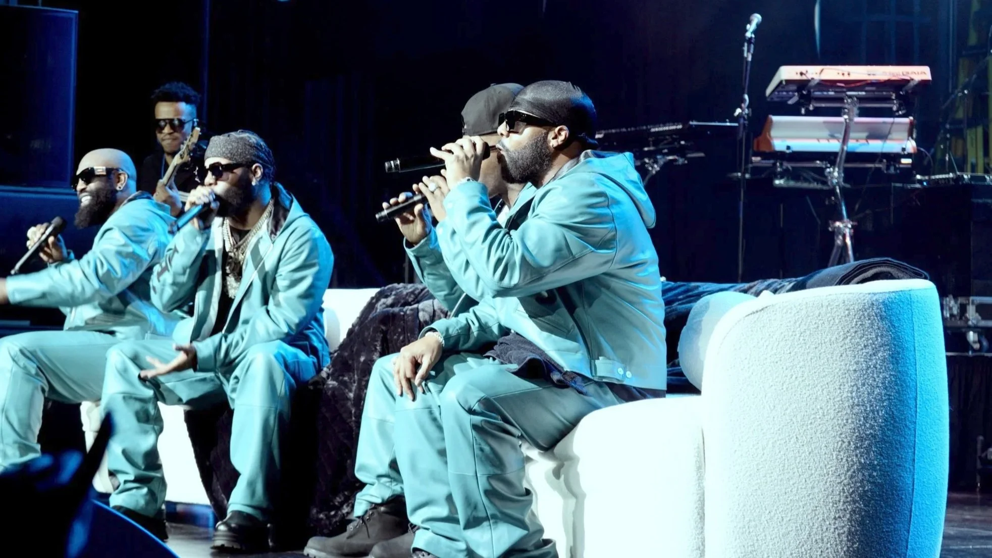 R&B group Jagged Edge performing on stage, wearing matching light blue outfits, with some holding microphones, and musical instruments visible in the background.