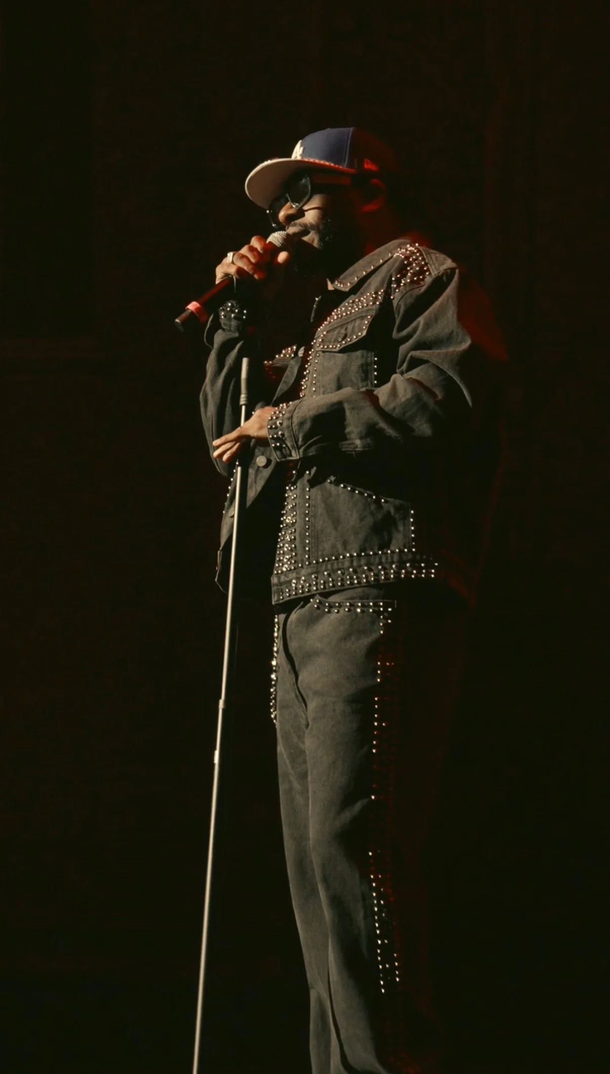 A man performing on stage with a microphone, wearing a denim jacket with metal stud embellishments, a baseball cap, sunglasses, and a dark outfit.