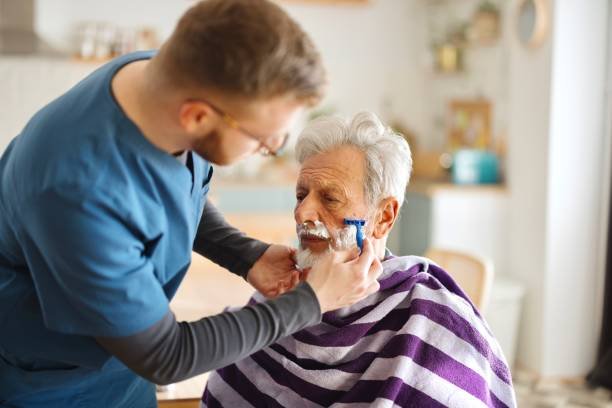 A young man giving a shave to an elderly man with a rotary razor in a cozy, well-lit room.