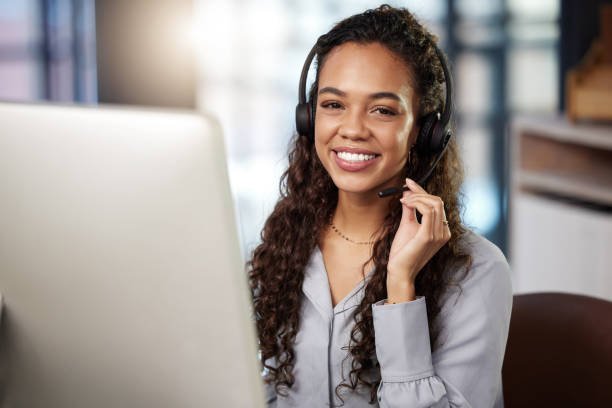 A woman with curly hair wearing a headset and smiling while working at a computer in an office setting.