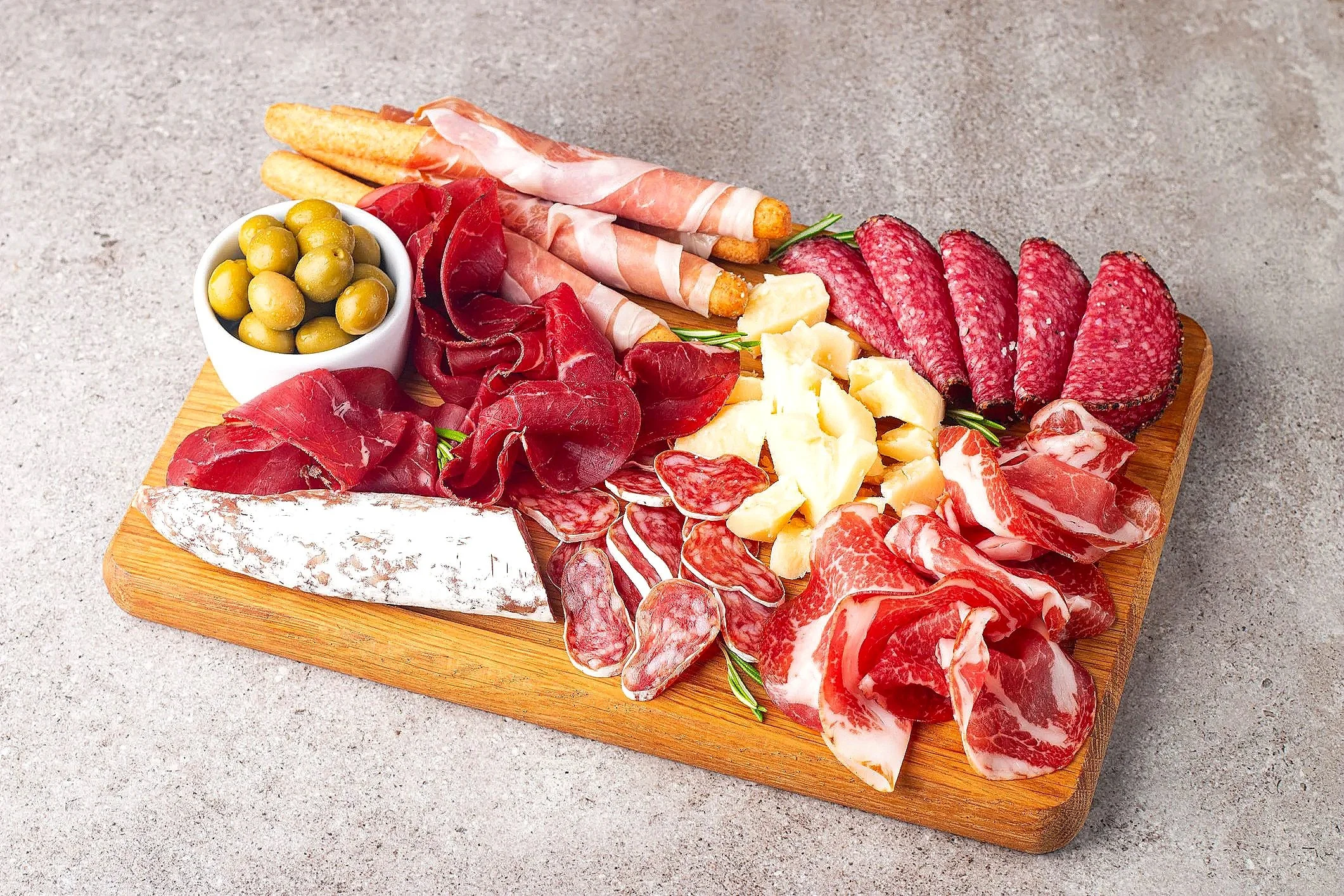 A wooden board with assorted cured meats, cheeses, green olives, and breadsticks.