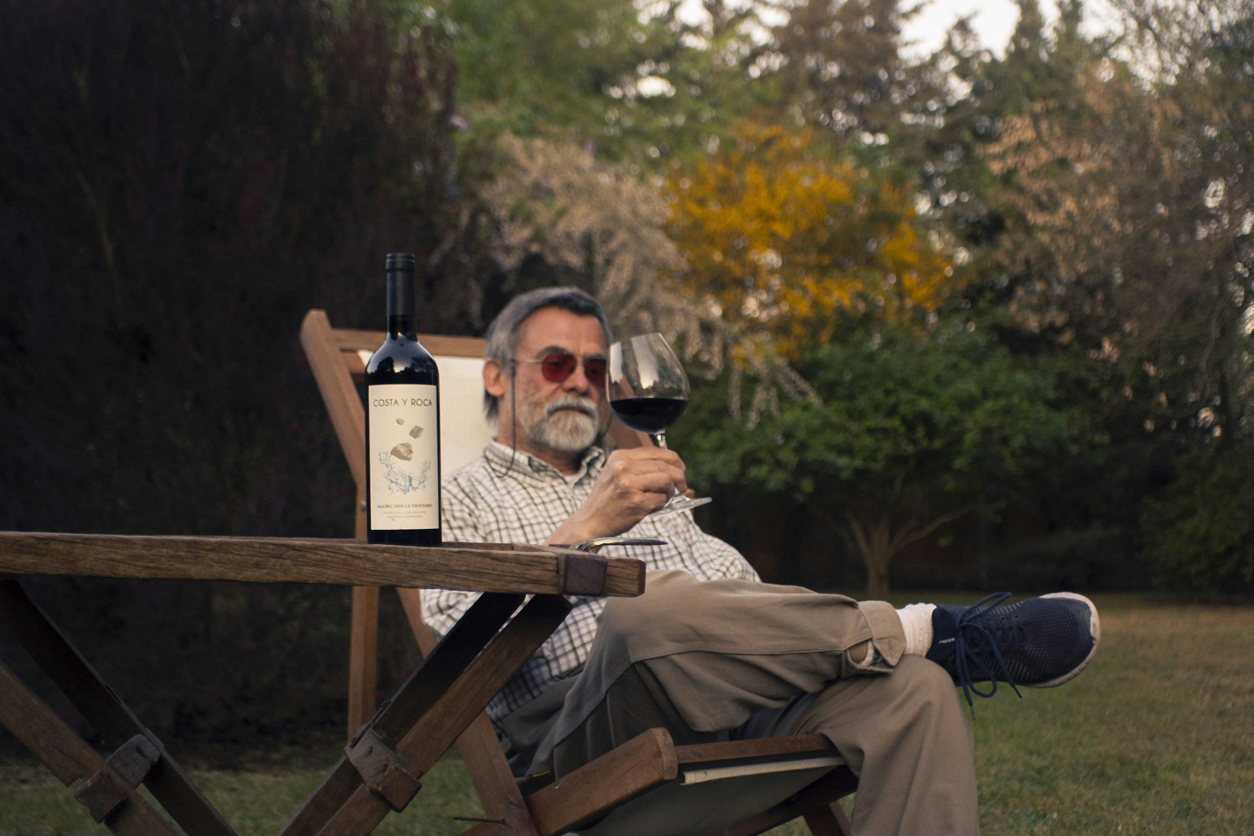 An older man with sunglasses sitting outside in a wooden chair, holding a glass of red wine, with a bottle of Costa y Roca wine on the table in front of him, surrounded by trees with autumn foliage.