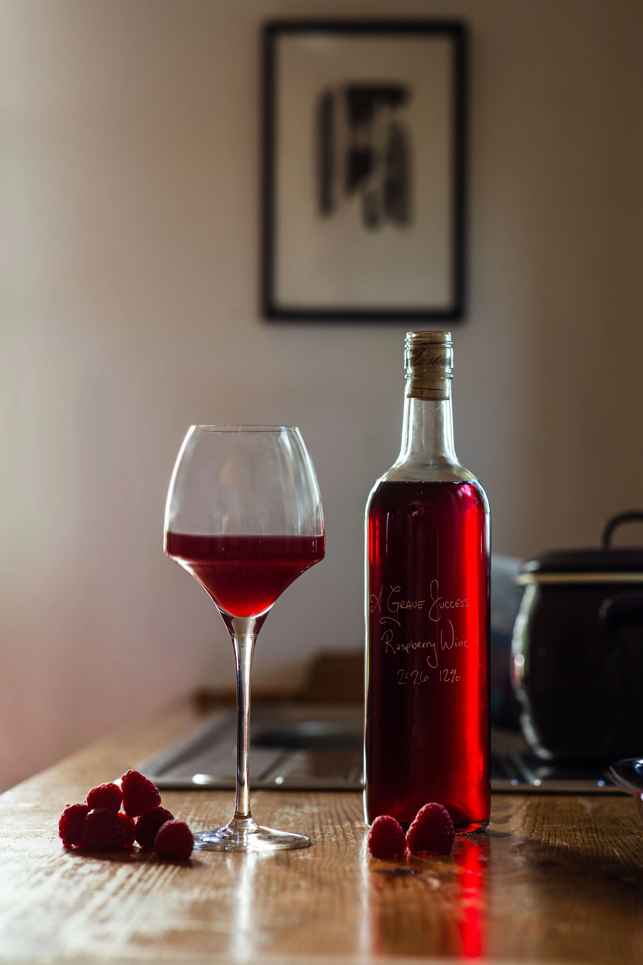A glass of raspberry wine and a bottle of raspberry wine sit on a countertop sprinkled with frersh raspberries