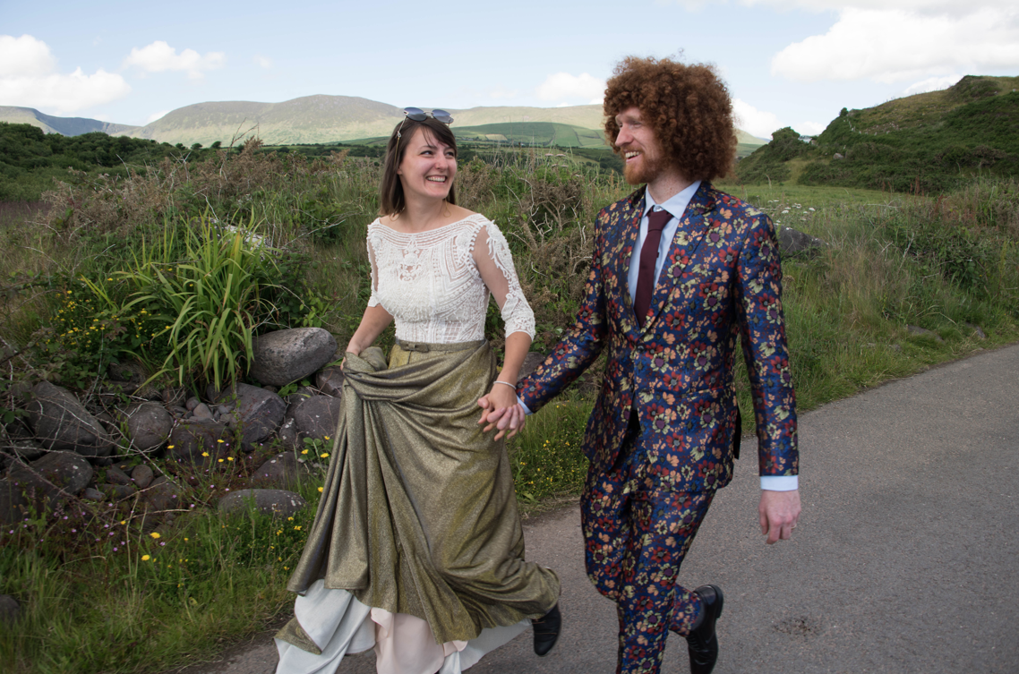 Winemakers Gretchen and Alan holding hands and walking along a countryside path surrounded by green hills and rocks, in their wedding outfits.