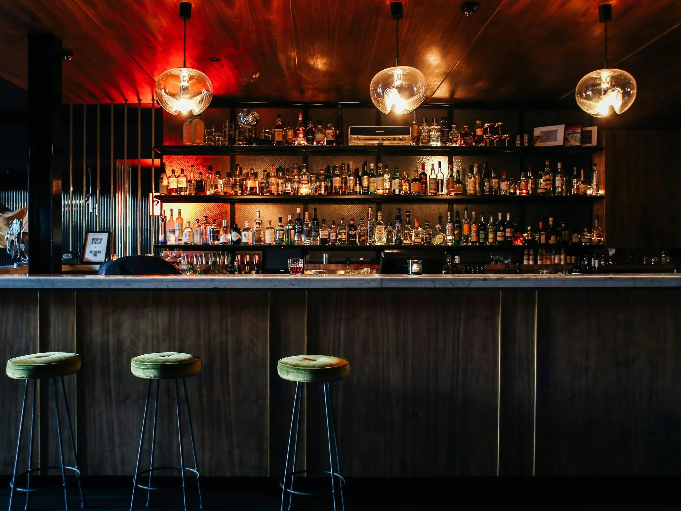 Interior of a dimly lit bar with a wooden ceiling and shelves stocked with various bottles of liquor. There are three green bar stools in front of the bar counter, and three pendant lights hanging from the ceiling.