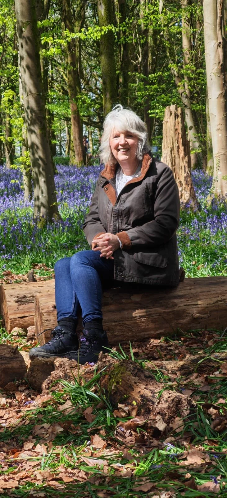 An woman with gray hair, wearing a brown jacket and blue jeans, sitting on a fallen log in a forest surrounded by green foliage and purple flowers.