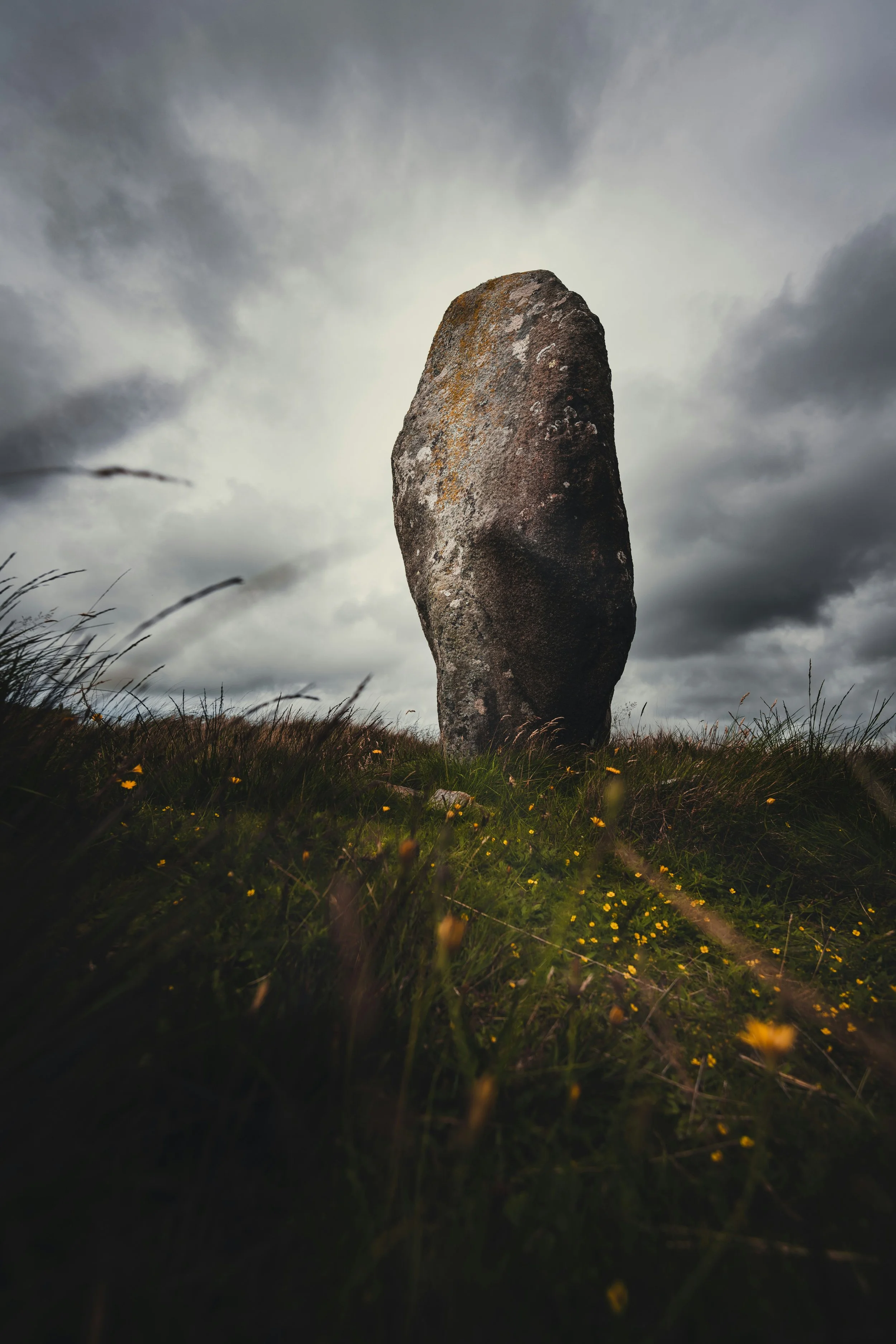 A large, upright standing stone or monolith on a grassy hill with yellow flowers, under a cloudy, overcast sky.
