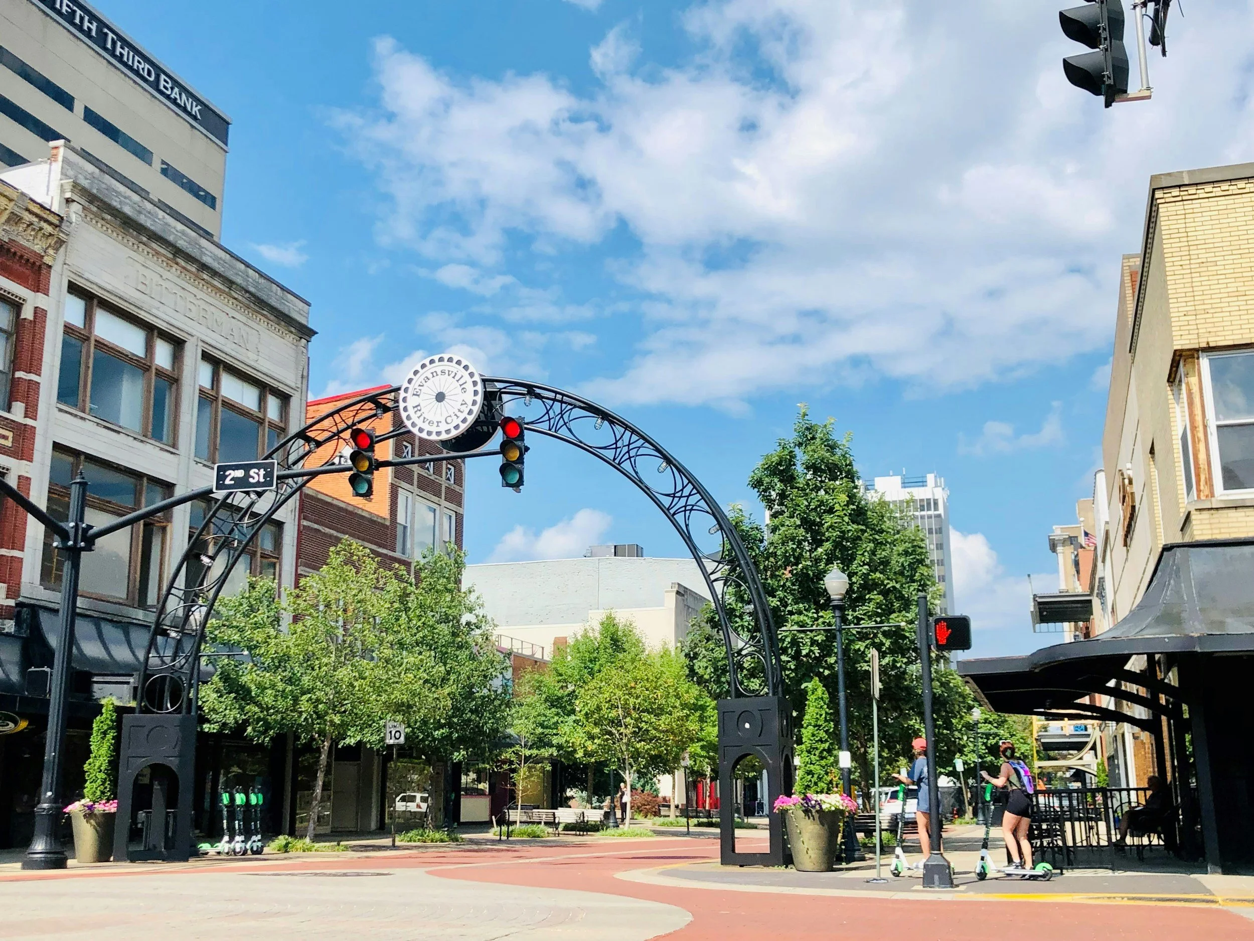 A city street scene with trees, buildings, and a decorative metal arch with a sign reading 'Evansville River City.' Traffic lights are hanging from the arch, with some red and green lights illuminated. Pedestrians with scooters are waiting to cross at a crosswalk, and a person is sitting on a bench under a black awning on the right side. The sky is partly cloudy.