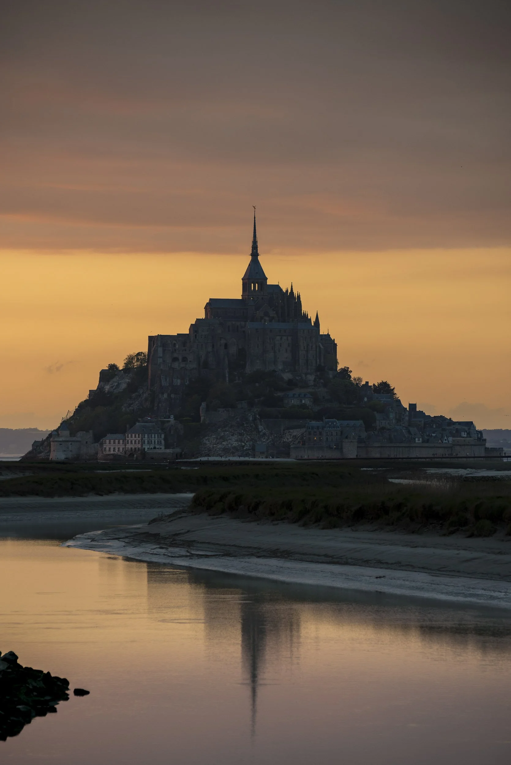 La imagen muestra una vista del Monte Saint-Michel al atardecer, rodeado de agua con reflejo de la estructura en la superficie.
