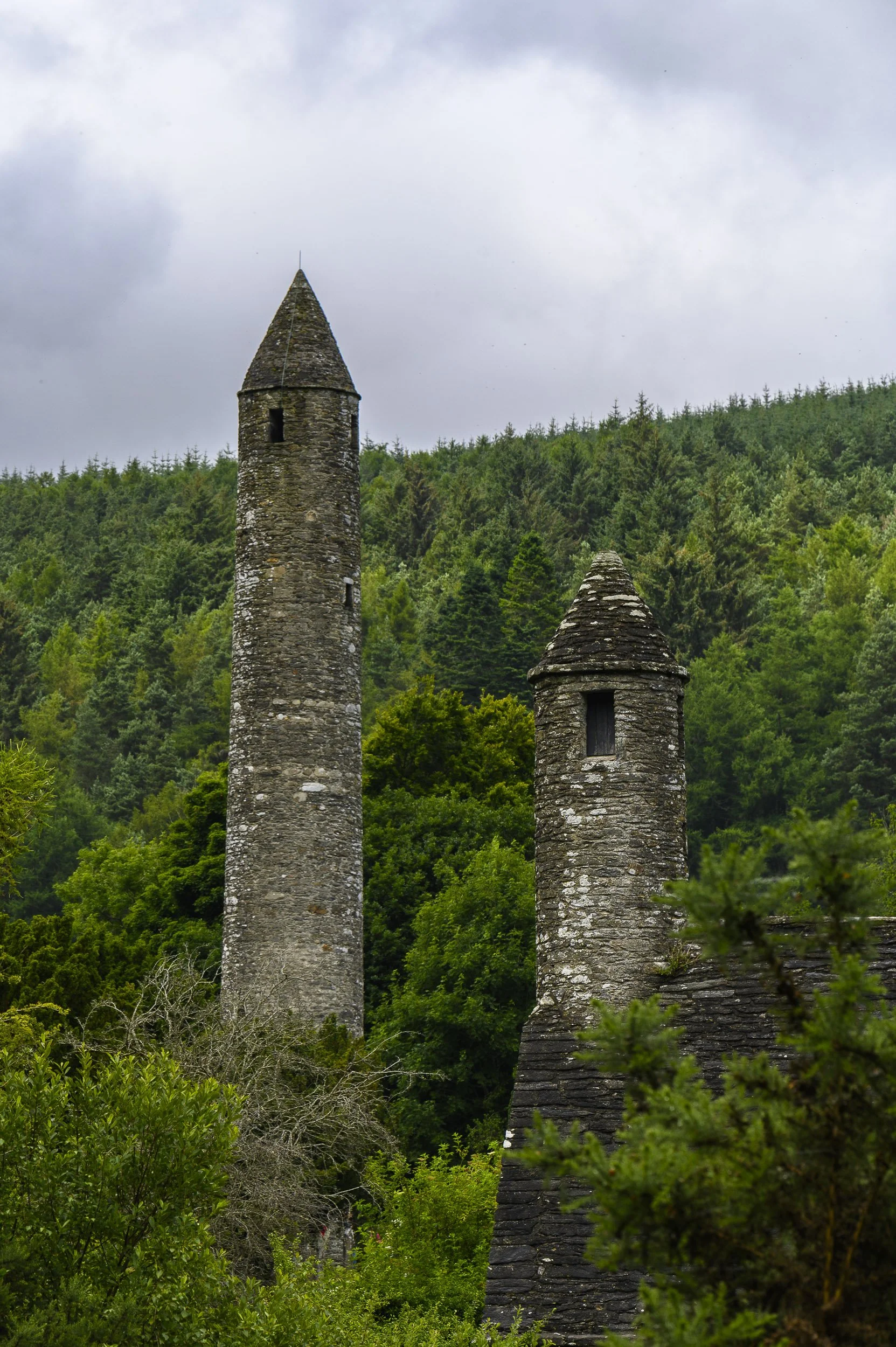 Dos torres de piedra en medio de un bosque verde en Glendalough, Irlanda, con nubes grises en el cielo.