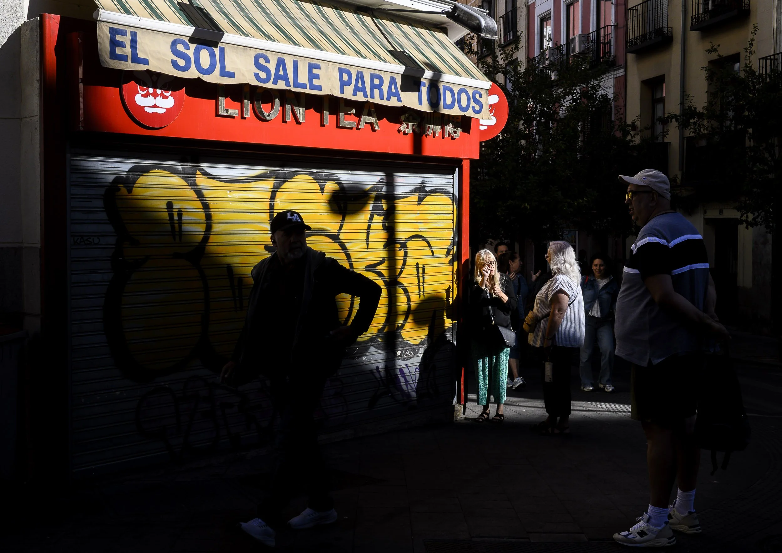 Personas en una calle cerca de un puesto con cartel que dice 'El sol sale para todos', en sombra y luz tenue.