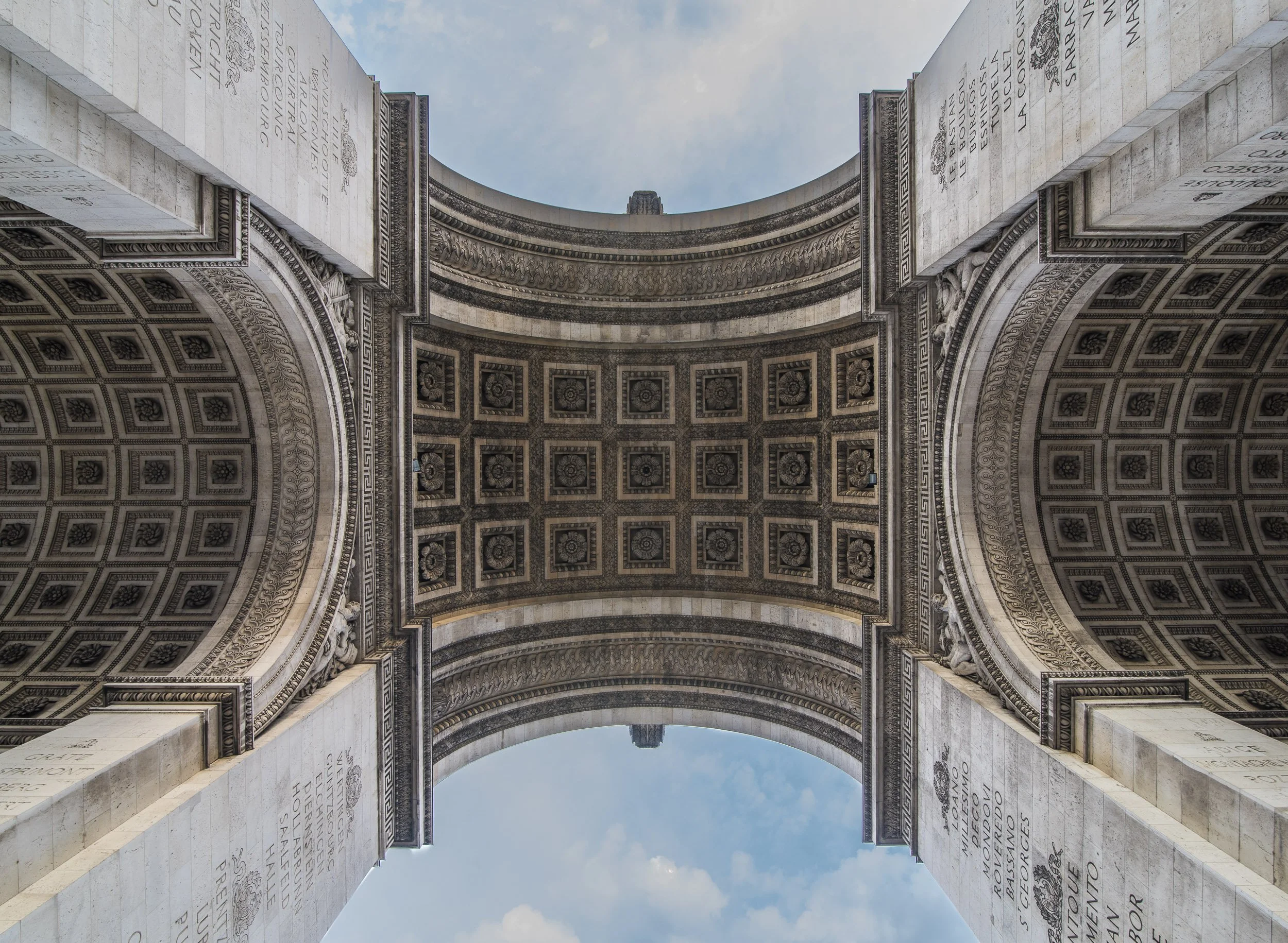 Vista desde abajo del Arco del Triunfo de París, con decoración detallada, inscripciones en columnas a los lados y cielo con nubes en el fondo.