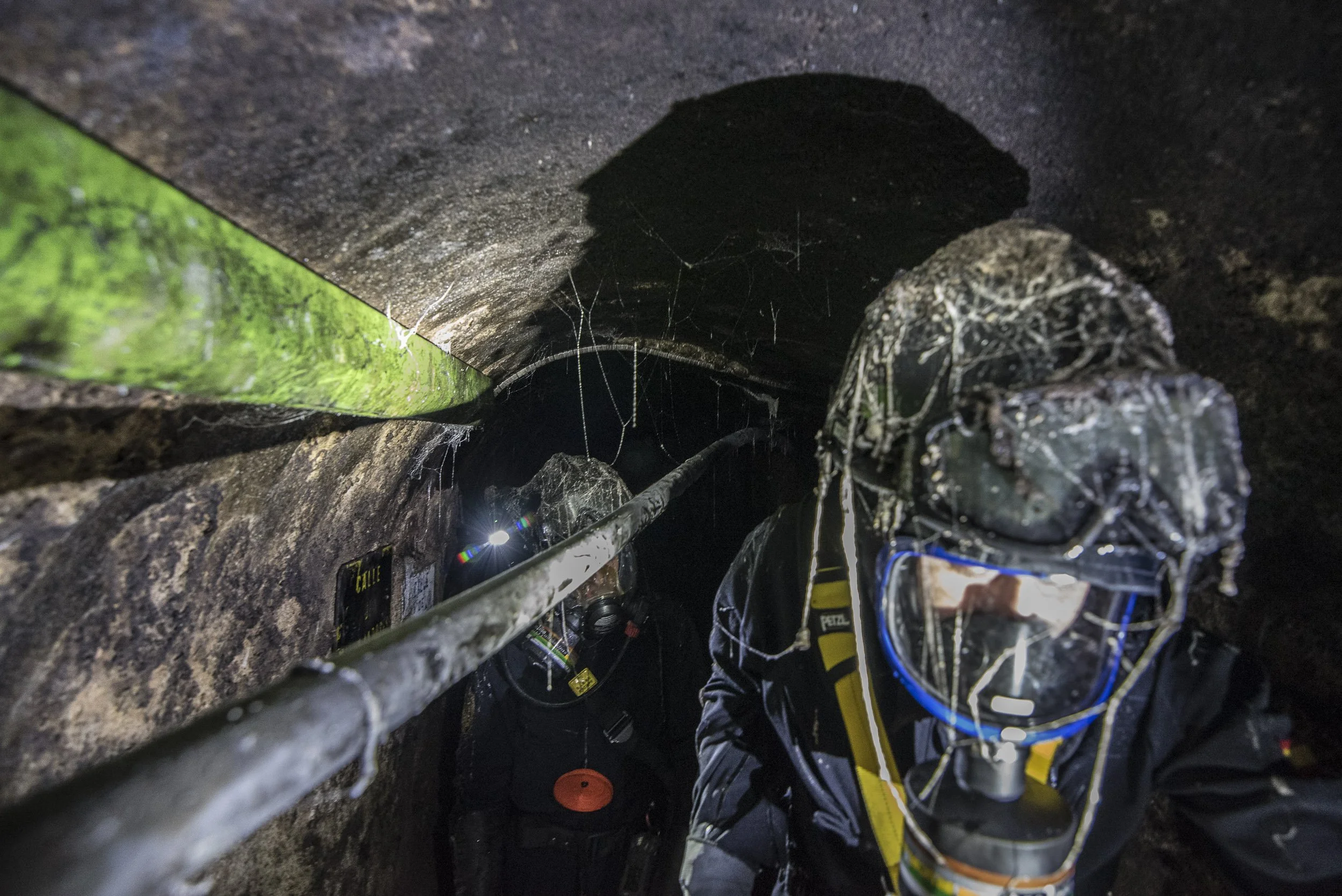 Mineros en una mina oscura con equipo de protección y cascos de seguridad, rodeados de telarañas y estructuras de excavación metálicas.