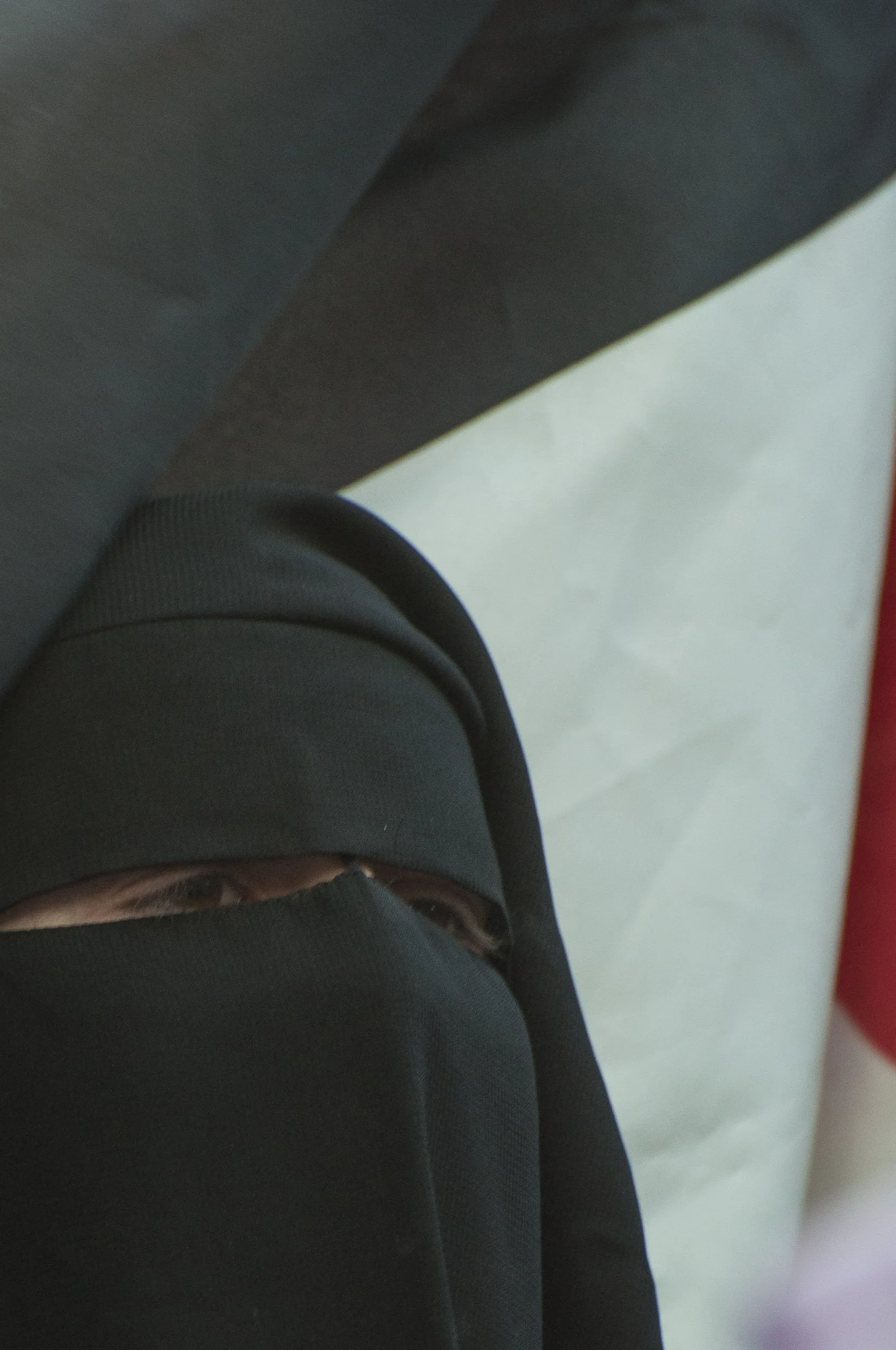 Una persona lleva un pañuelo negro que cubre la mayor parte de la cara, dejando ver solamente los ojos, con un fondo neutro y superficie de suelo blanca.