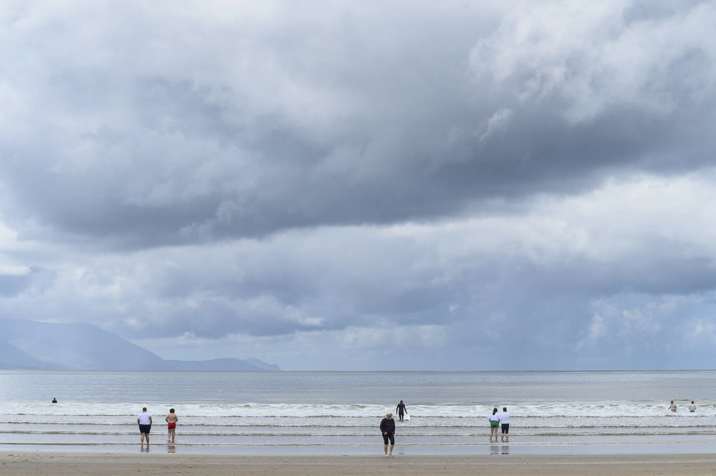 Playa con varias personas disfrutando en la orilla, con cielos nublados y montañas en la distancia.