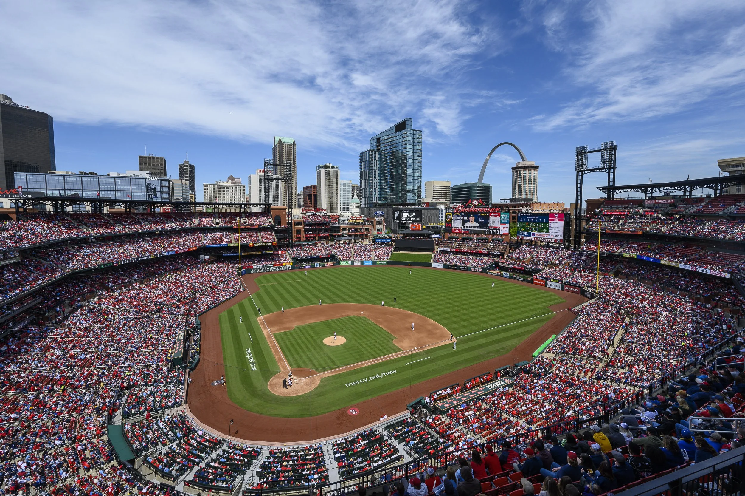 Vista panorámica de un campo de béisbol en un estadio lleno de espectadores, con una ciudad moderna de rascacielos y el arco de la ciudad en el fondo bajo un cielo parcialmente nublado.