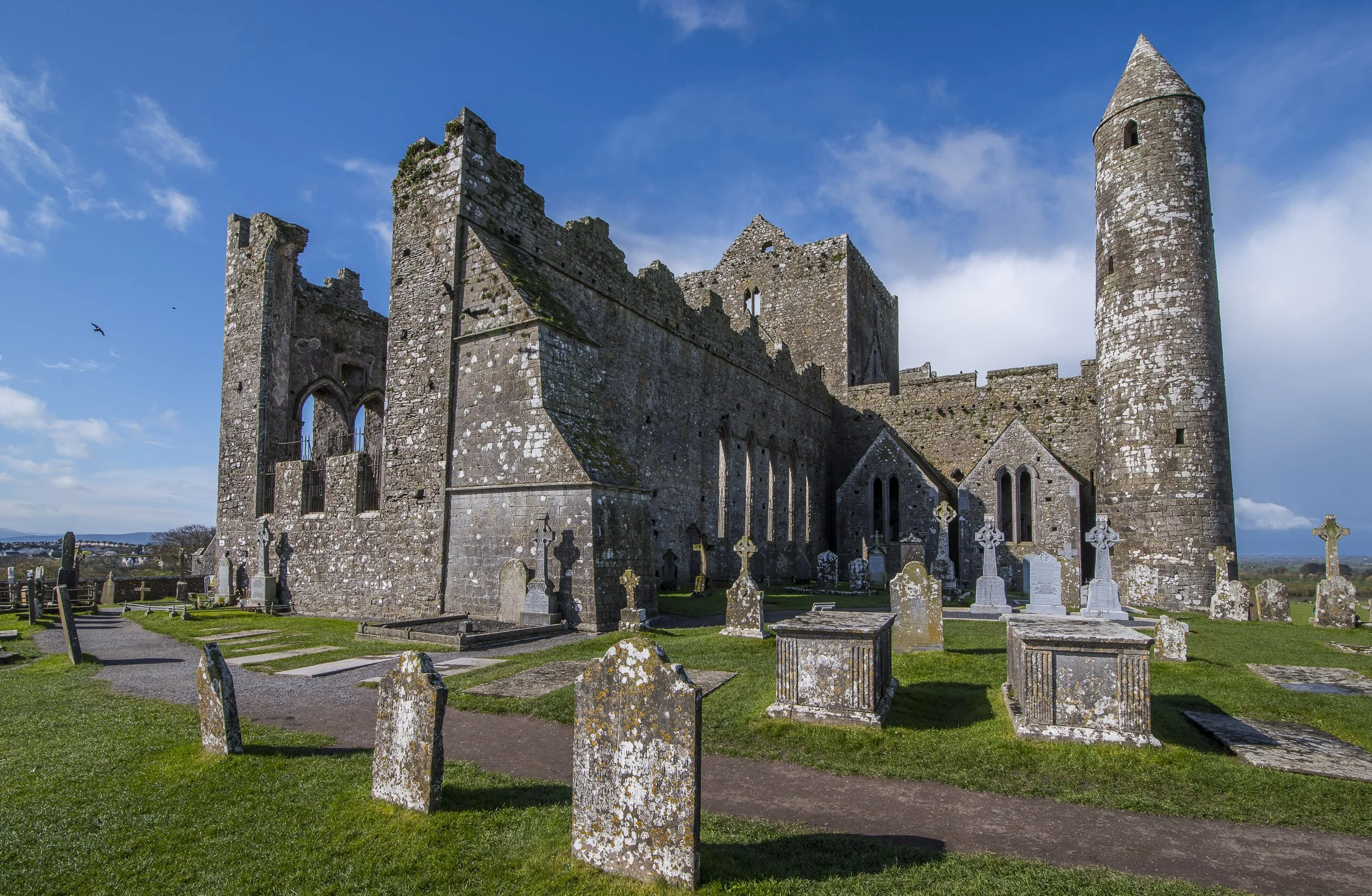 Ruinas de la Roca de Cashel en Irlanda, con muros de piedra y una torre en un día soleado, rodeado de tumbas y césped verde.