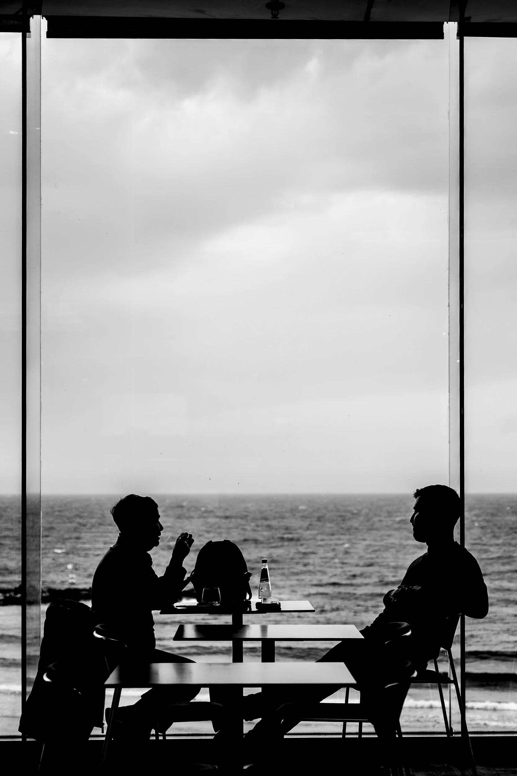 Dos personas conversando en un café con vista al mar en la playa de Tel Aviv, en silueta, en un día nublado.